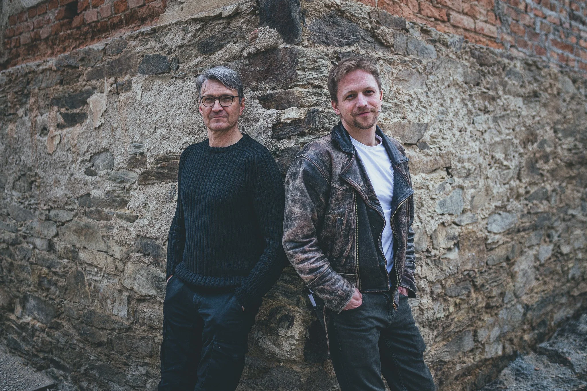 Two men standing against a stone and brick wall, with casual attire, looking at the camera.