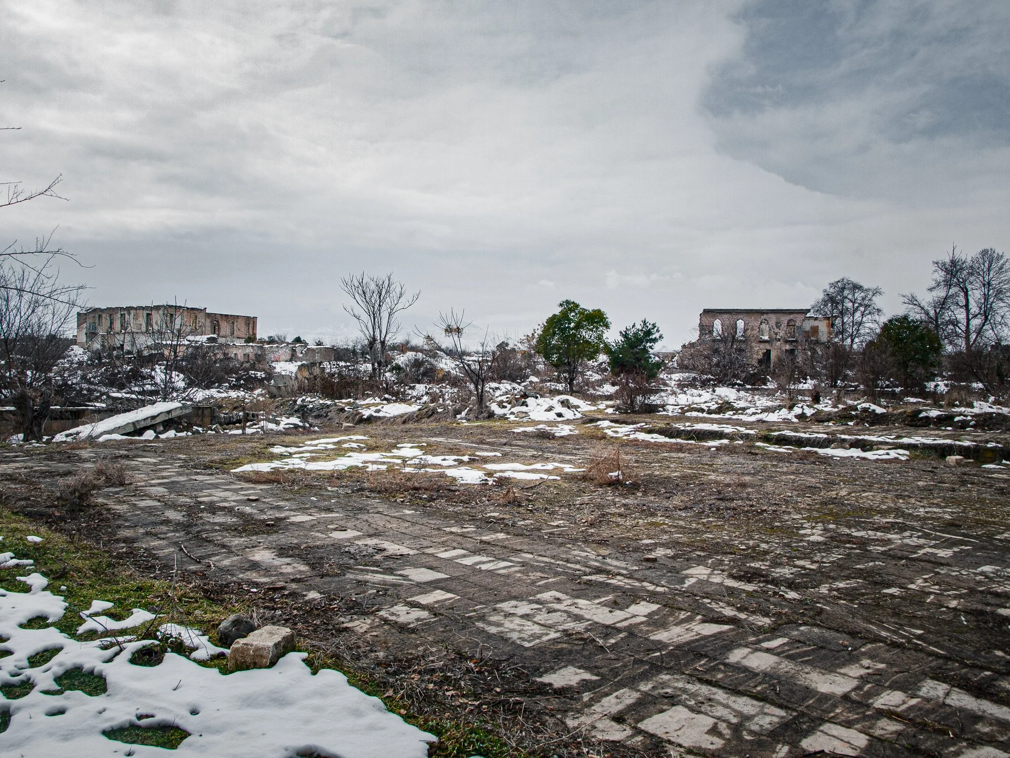 Ruined buildings and leafless trees in a snowy, desolate landscape with a cloudy sky.