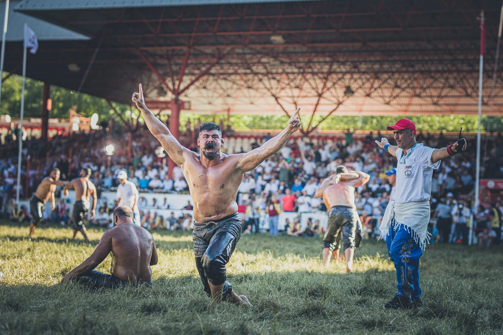 A man celebrating on a grassy field during a traditional yurt festival, with an excited expression, arm raised, and crowd in the background.