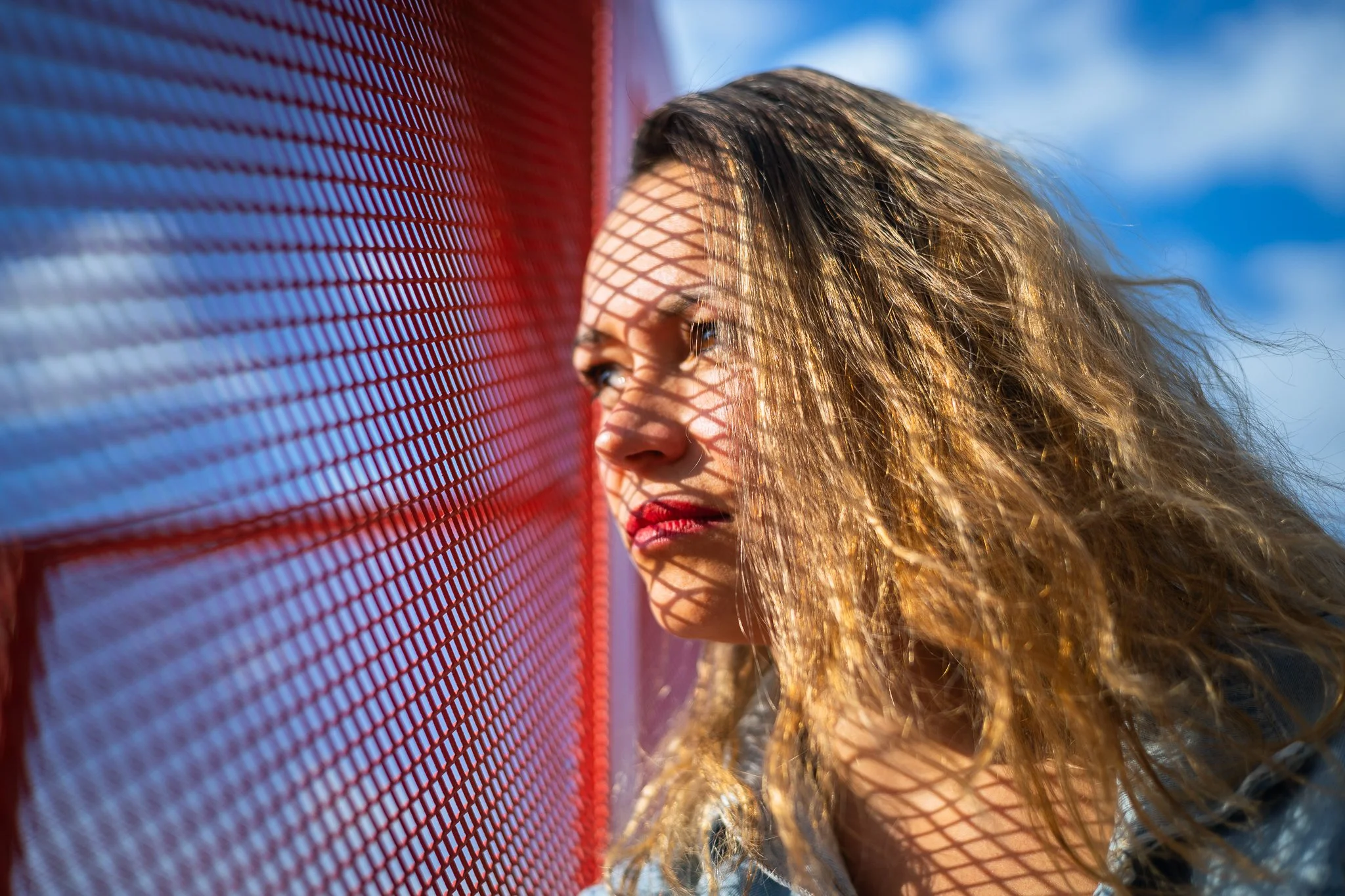 A woman with curly hair and red lipstick looks through a red fence, with her face casting a shadow of the fence's pattern.