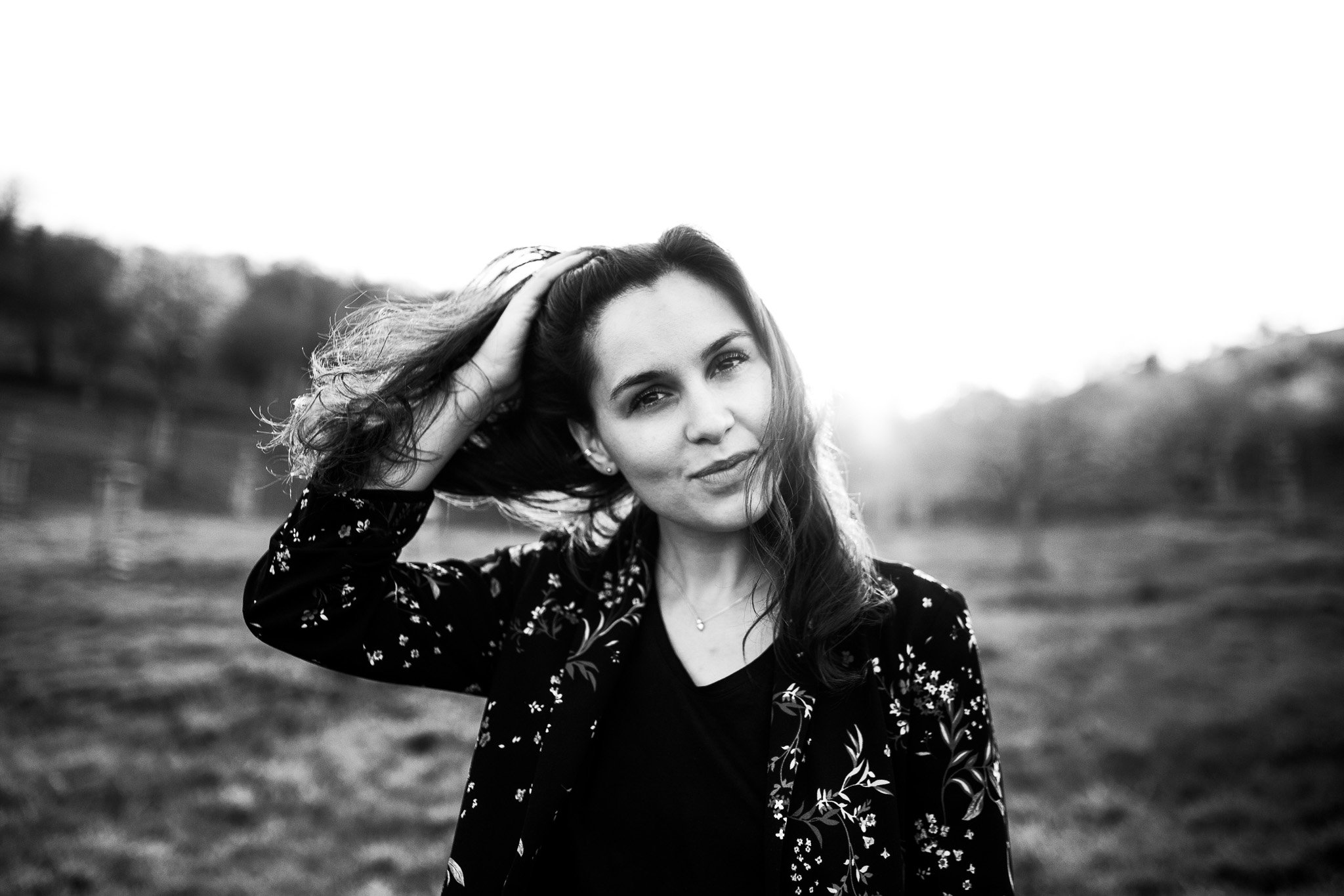 Black-and-white photo of a woman outdoors, gazing at the camera, with one hand in her hair, wearing a dark top and a floral blazer, with a blurred natural background.