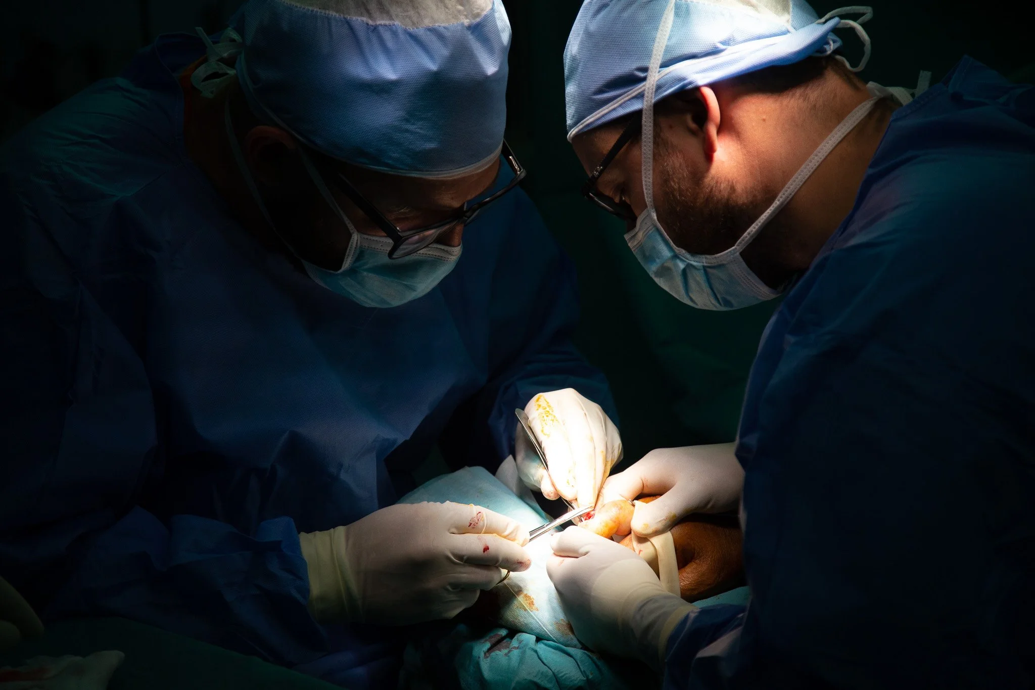 Two surgeons in scrubs and masks performing surgery under bright operating room lights.