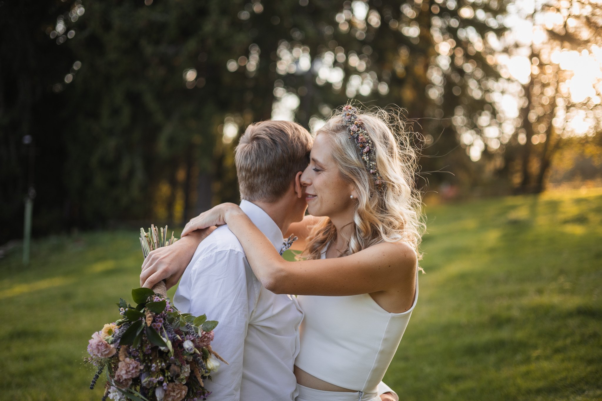 A smiling woman and man embrace outdoors on a sunny day, woman holding a bouquet of flowers, trees in the background.