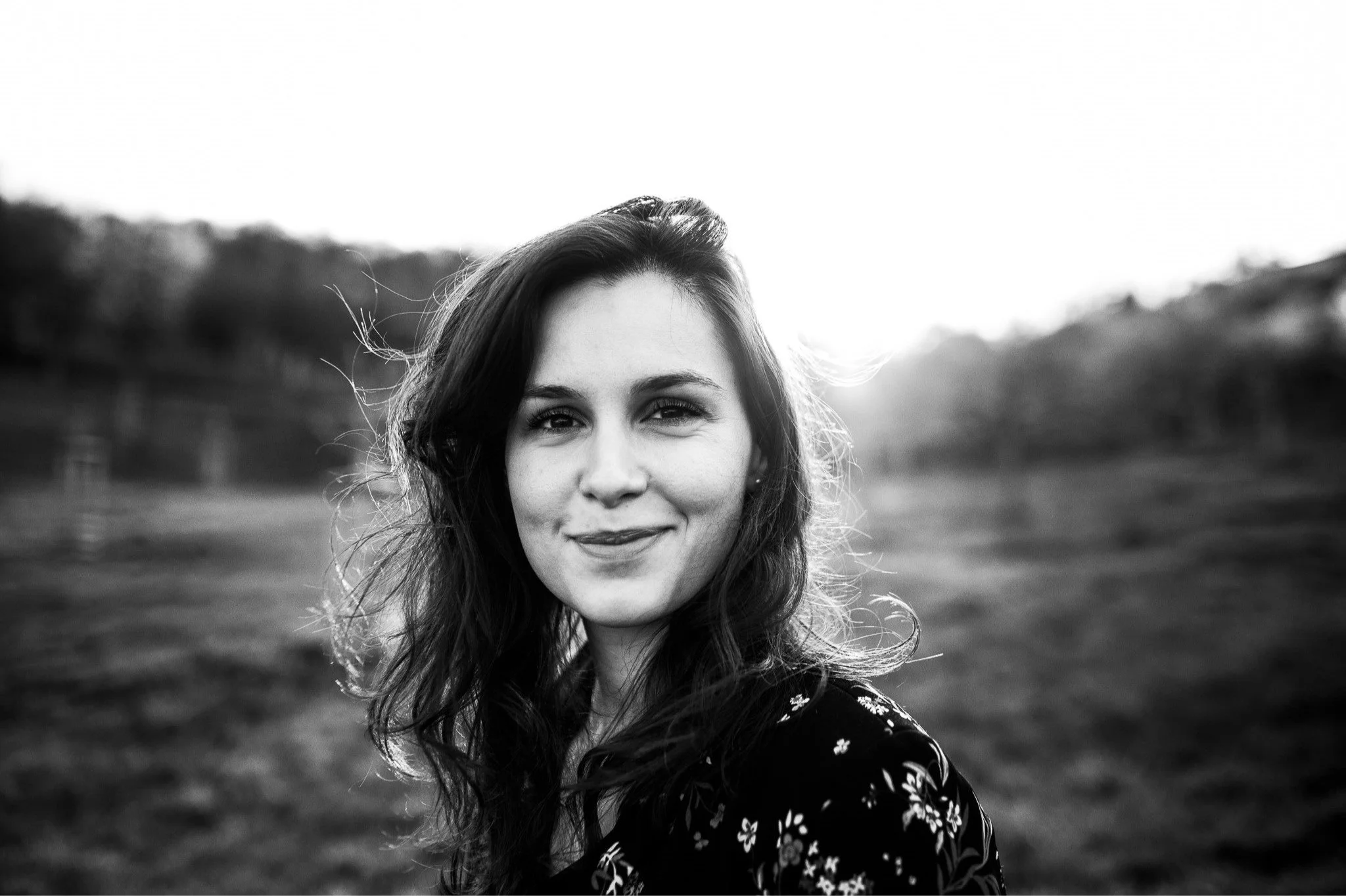 A black and white photo of a young woman with wavy hair outdoors with trees in the background, smiling at the camera.