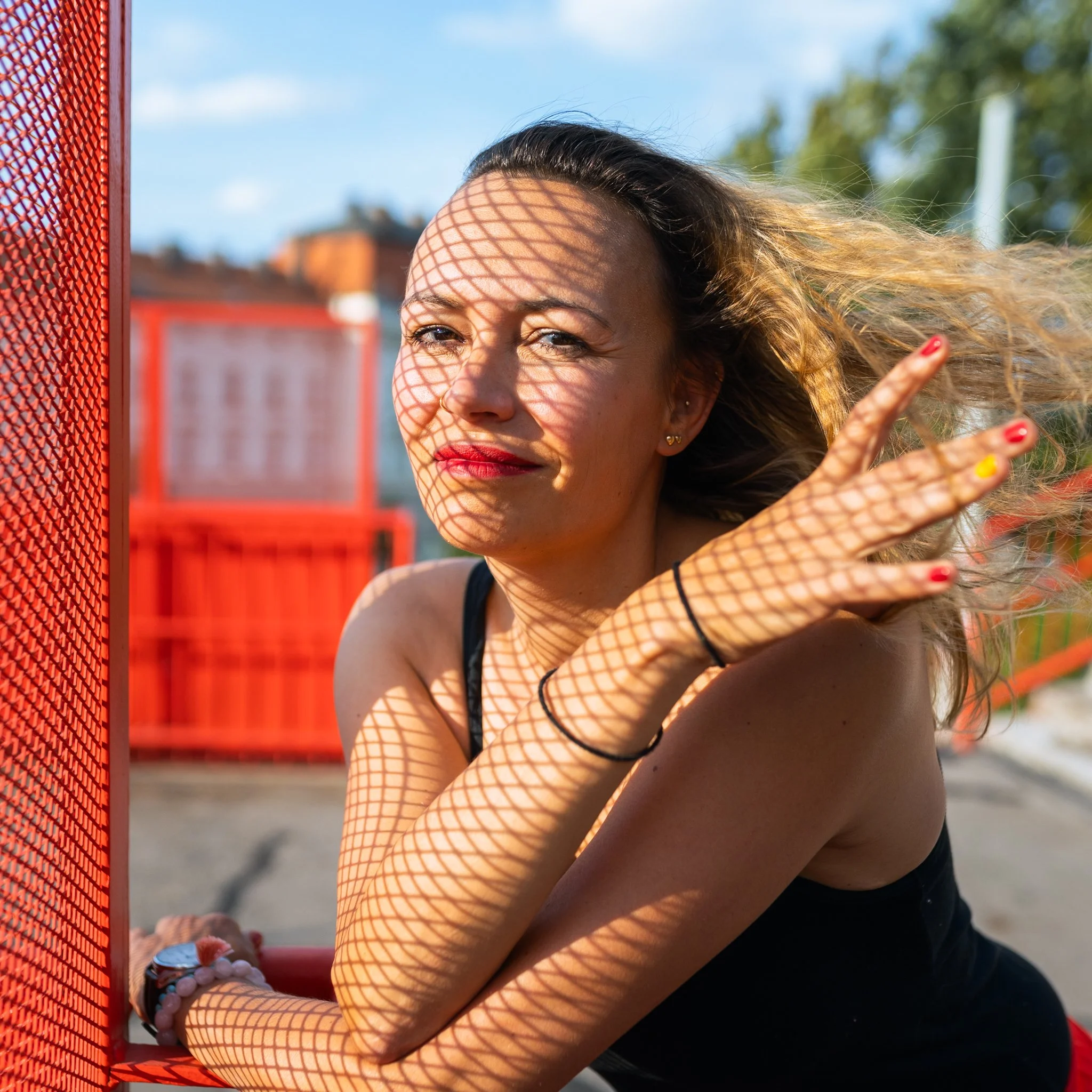 A woman with blonde hair and red lipstick posing outdoors behind a red metal fence, casting a shadow pattern on her face with the sunlight.
