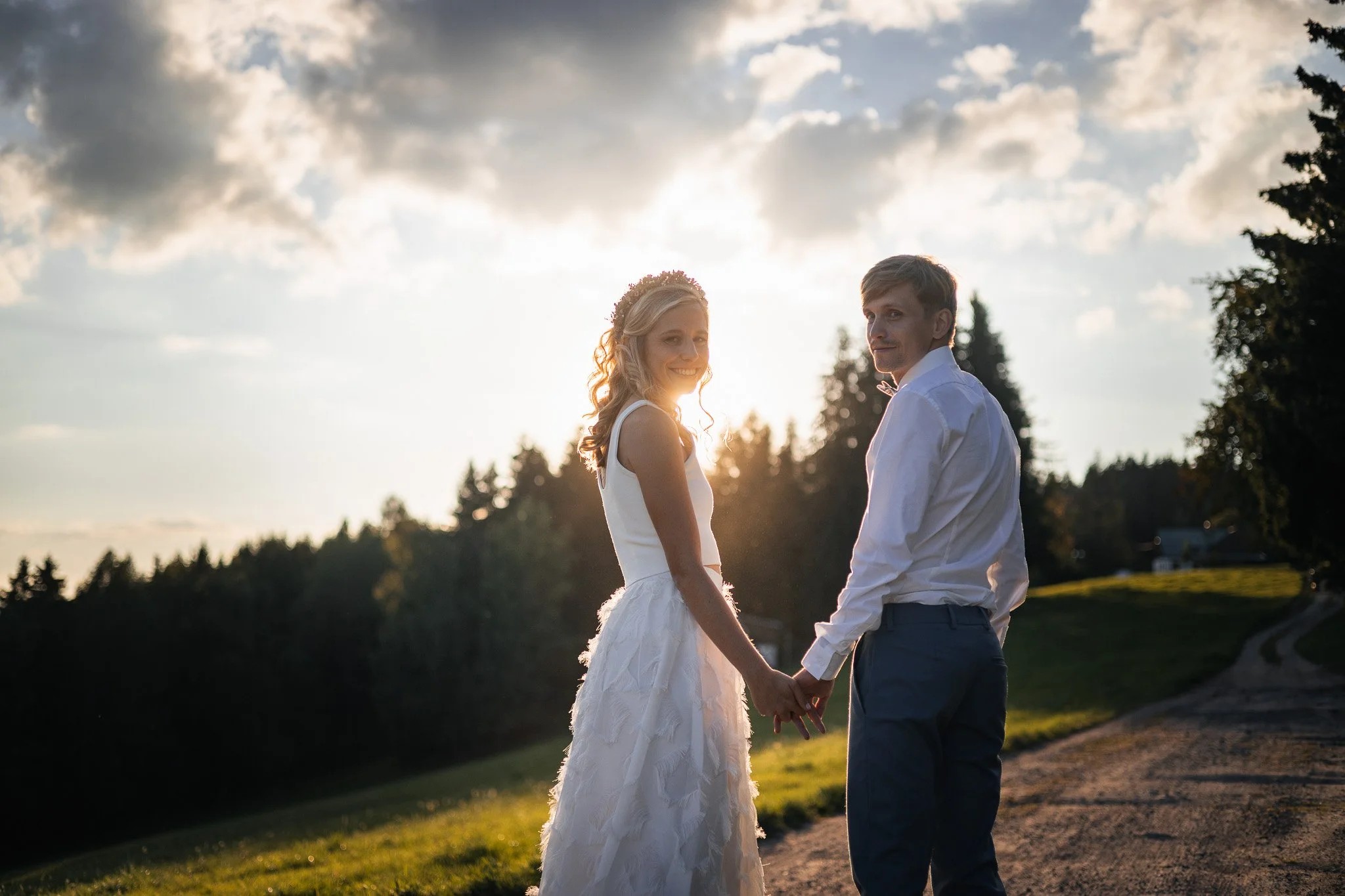 A bride and groom holding hands outdoors during sunset, with the bride smiling and wearing a white wedding dress and floral crown, and the groom dressed in a white shirt and dark pants.