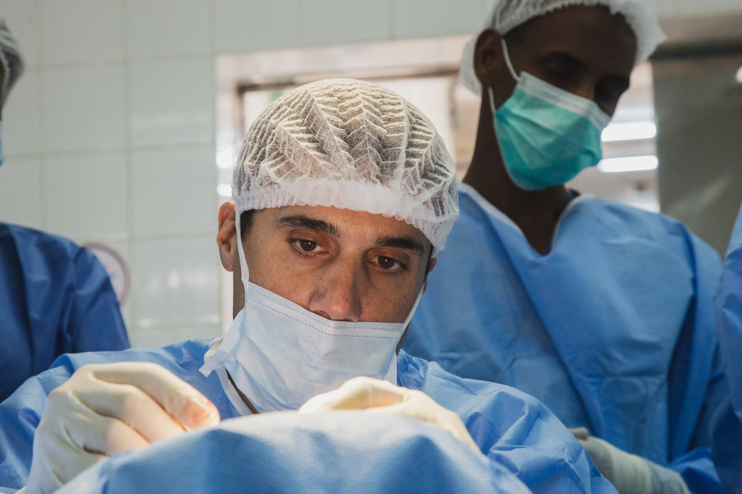 Two surgeons in operating room, one focusing on a surgical procedure, both wearing masks, gloves, and surgical caps.