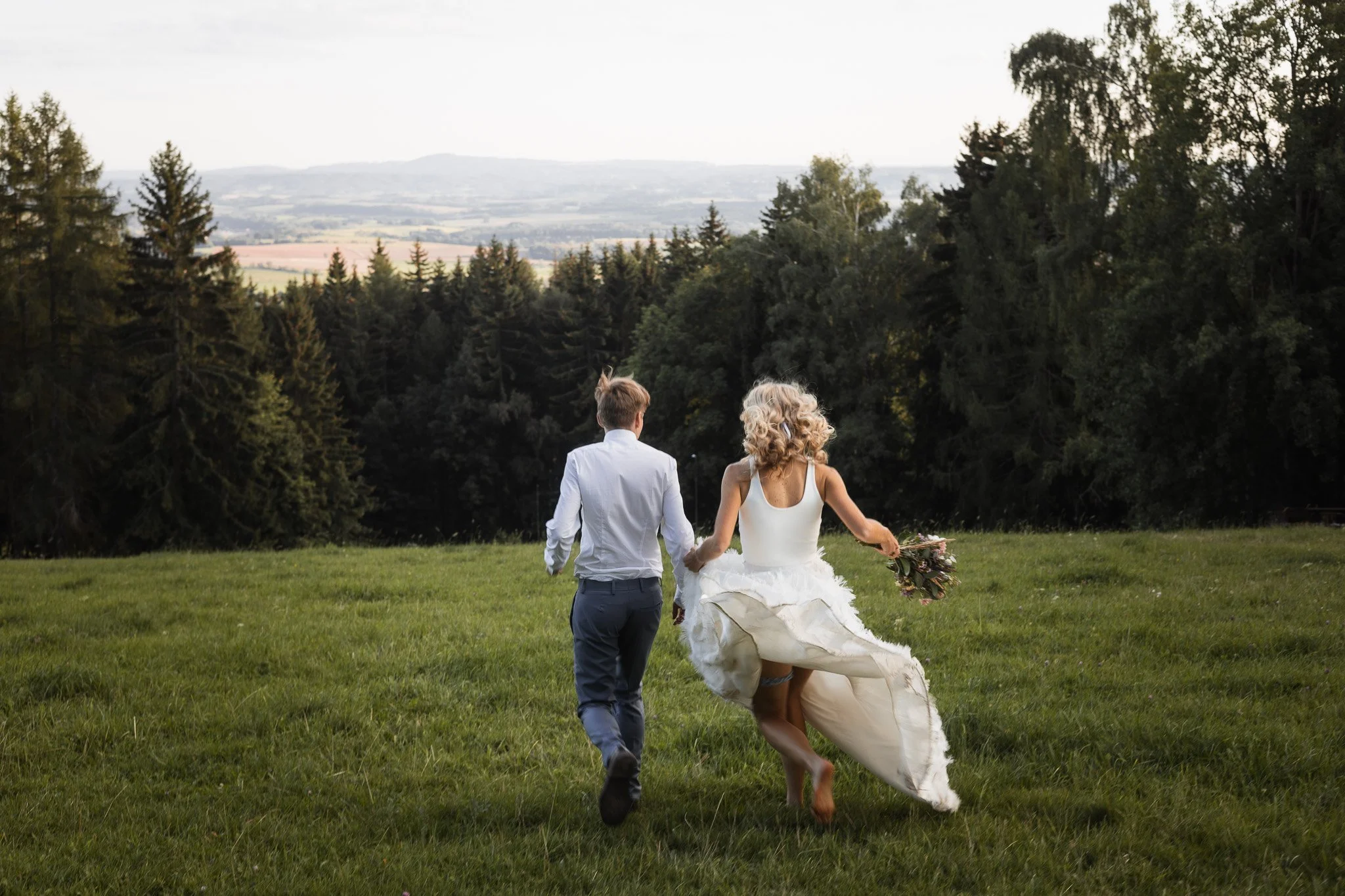 A newlywed couple runs across a grassy field, with the bride holding a bouquet, surrounded by trees with a distant landscape in the background.