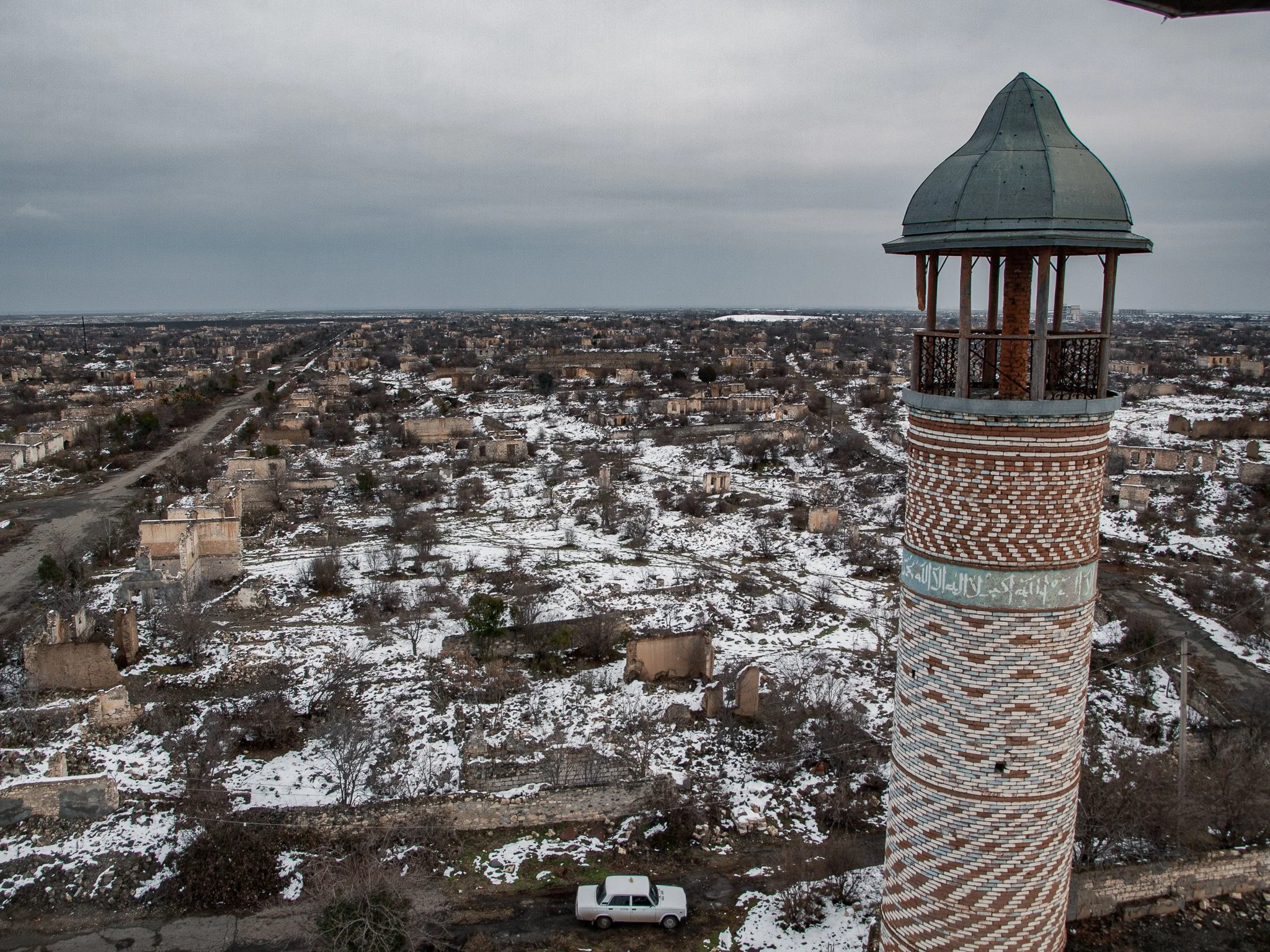 A brick minaret with a green domed roof overlooking a snowy, abandoned landscape with scattered ruins and trees under a cloudy sky.
