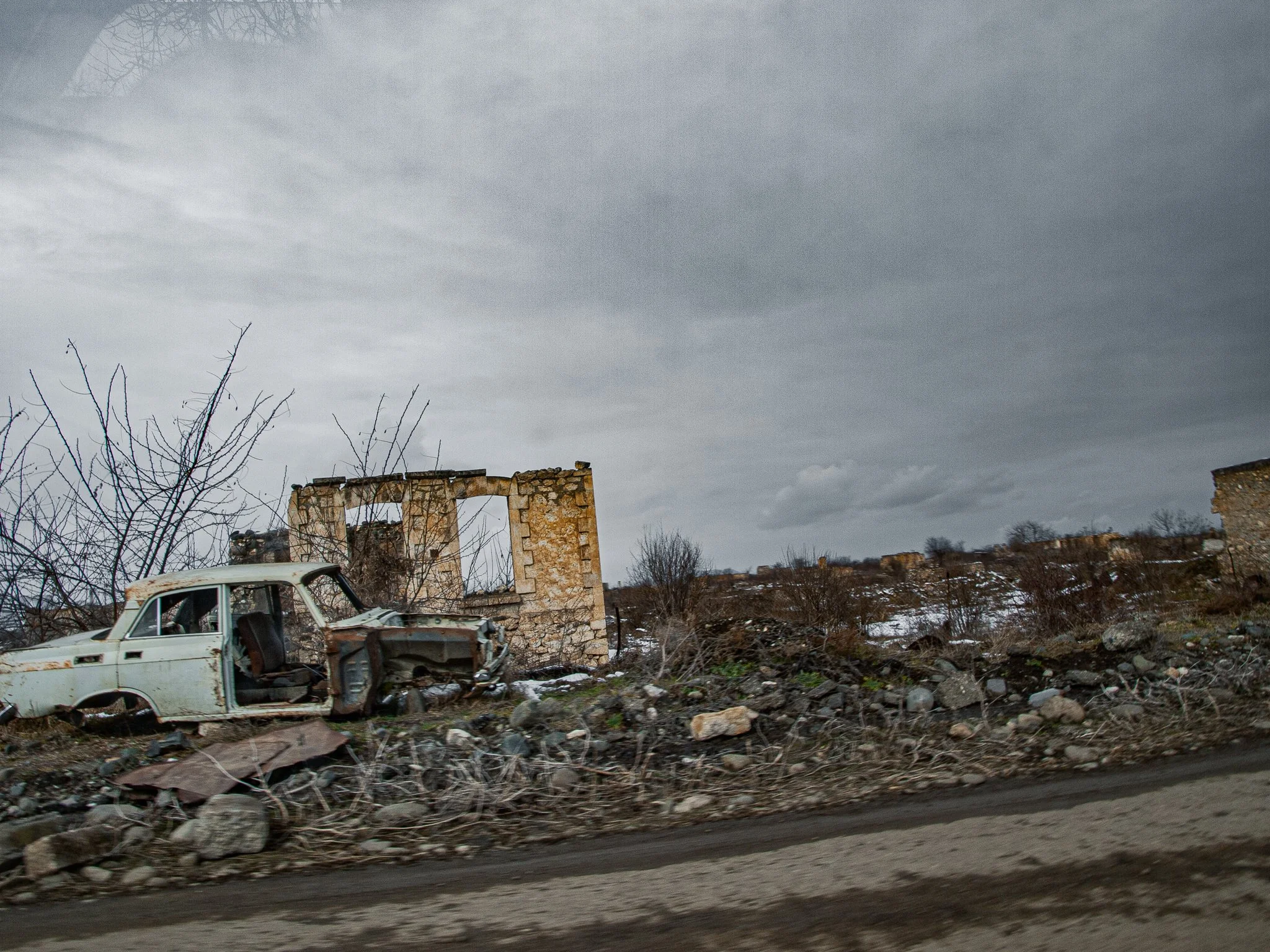 Abandoned, dilapidated car and ruined building in a desolate, rocky landscape under a cloudy sky.