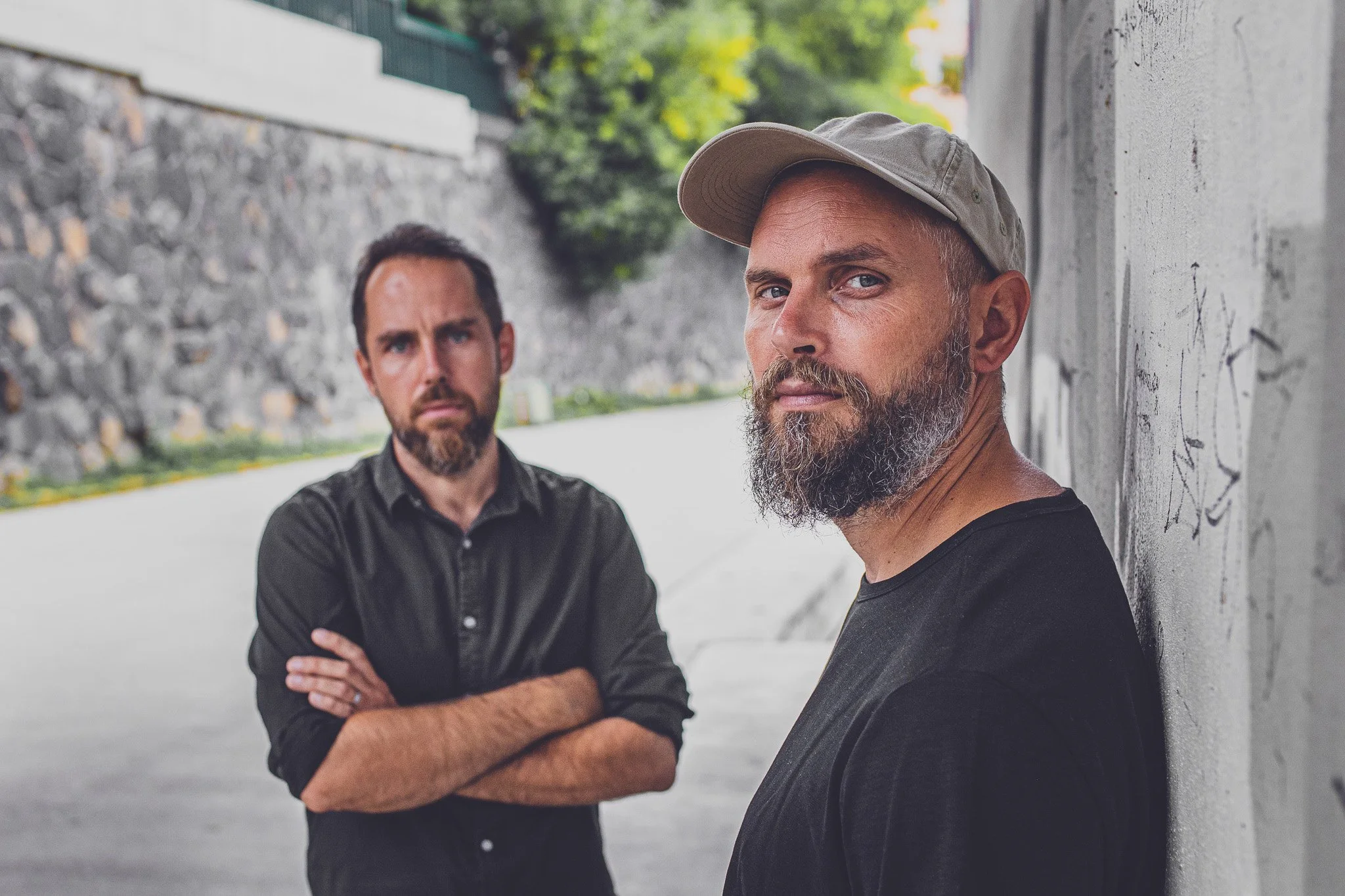 Two men standing outdoors, one with a beard and baseball cap leaning against a wall, the other with arms crossed and looking serious in the background.