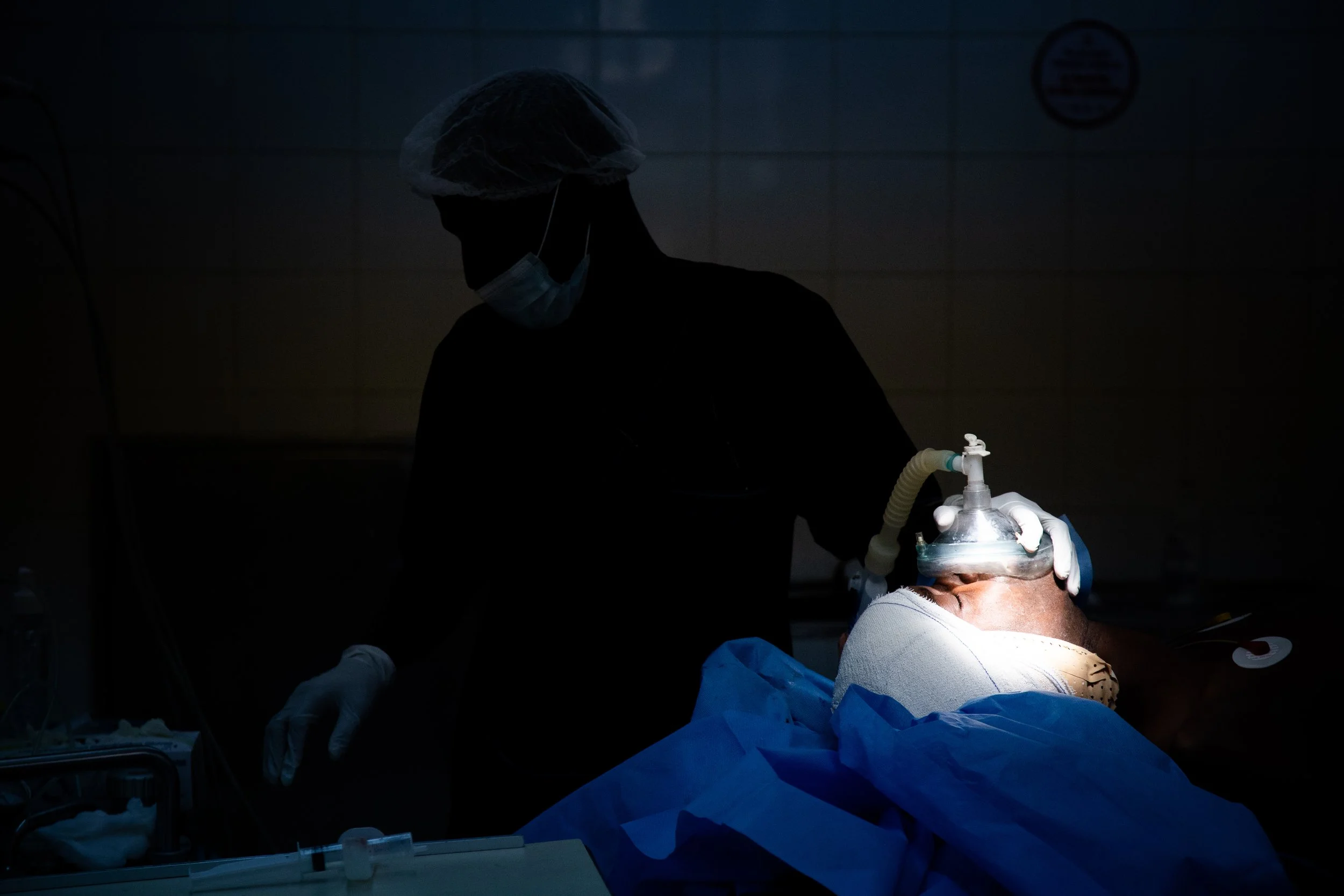 A healthcare professional in scrubs, mask, gloves, and hair cover performing a medical procedure on a patient with a ventilator mask.