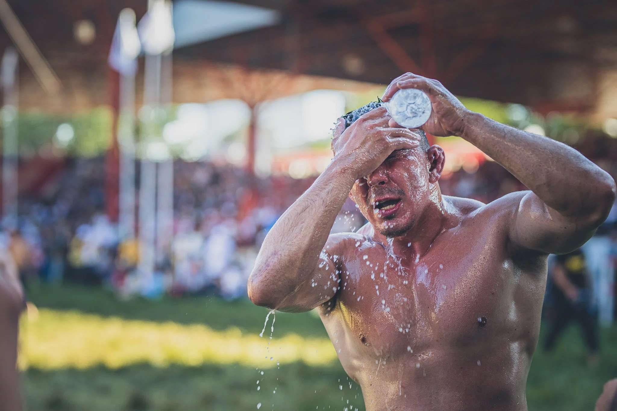 A shirtless man with wet hair and skin, pouring water on his head during an outdoor event under a shaded structure, with a blurred crowd in the background.
