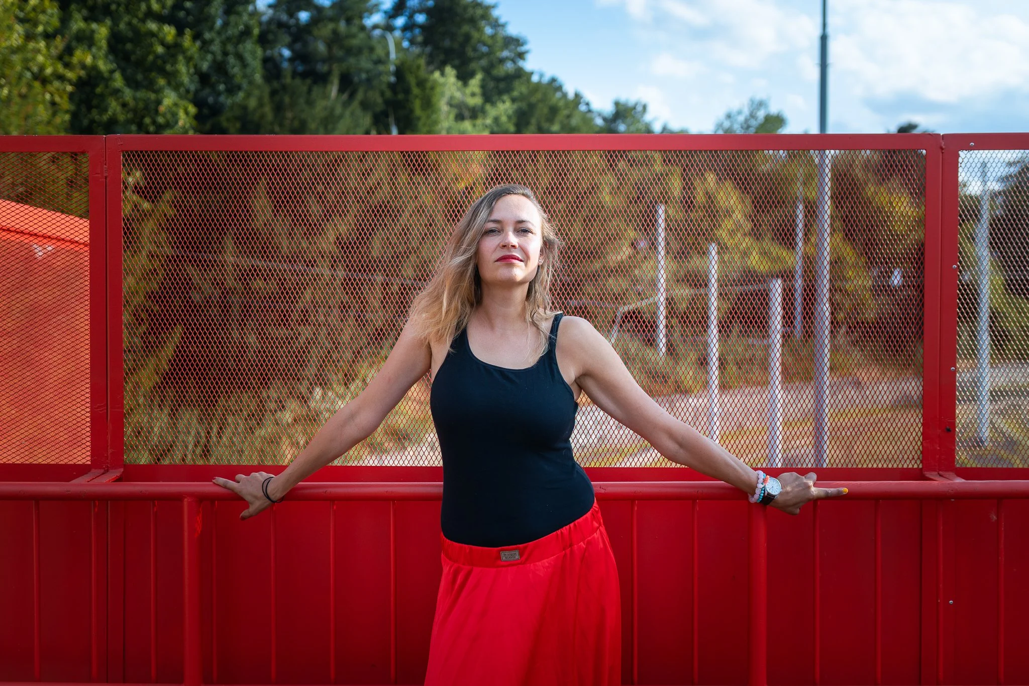 A woman in black tank top and red pants stands outdoors, leaning on a red metal fence with green trees and a partly cloudy sky in the background.