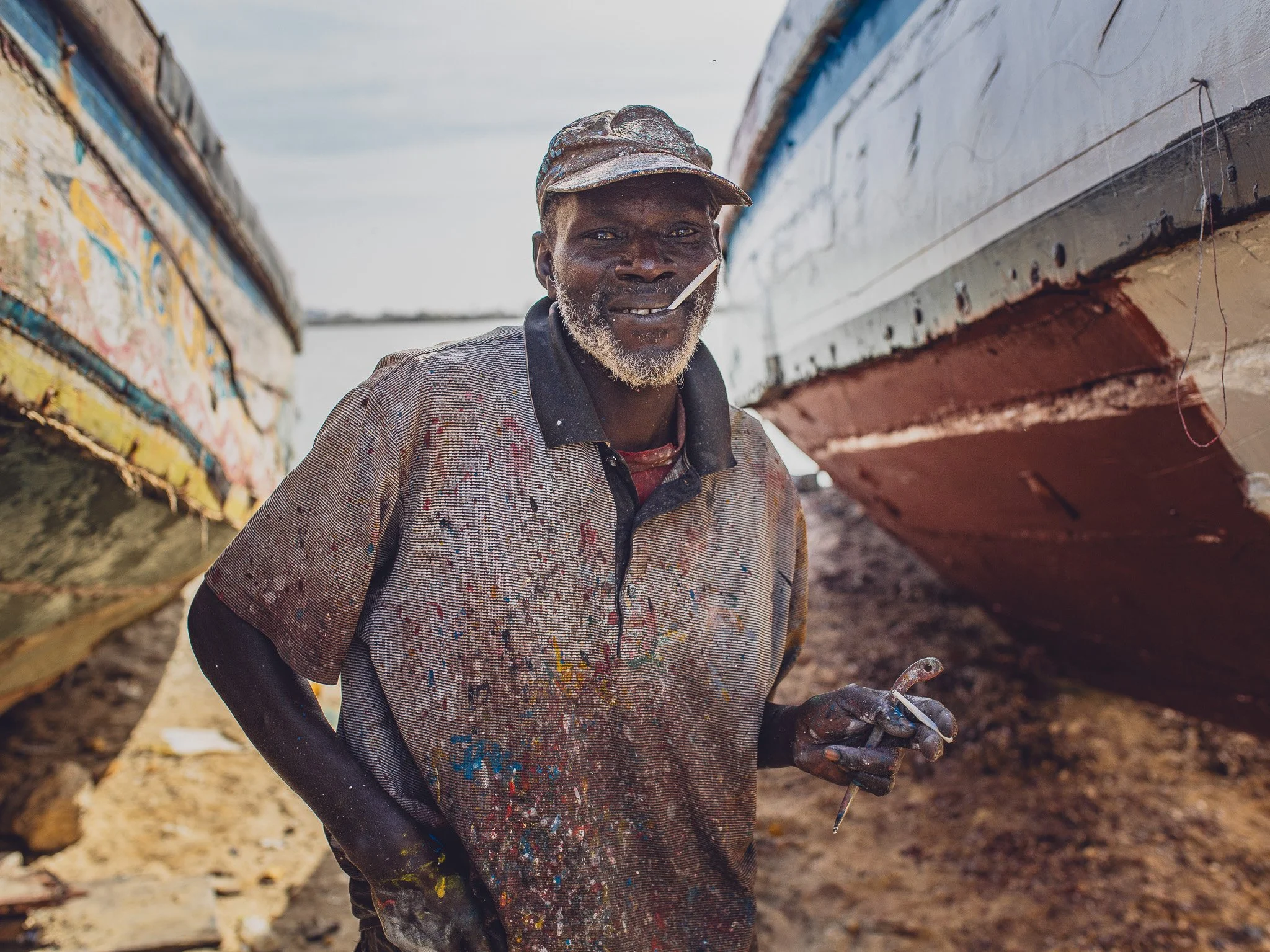 A man standing between two painted boats, holding a paintbrush, wearing a splattered shirt and a cap, with a beard and a toothpick in his mouth.