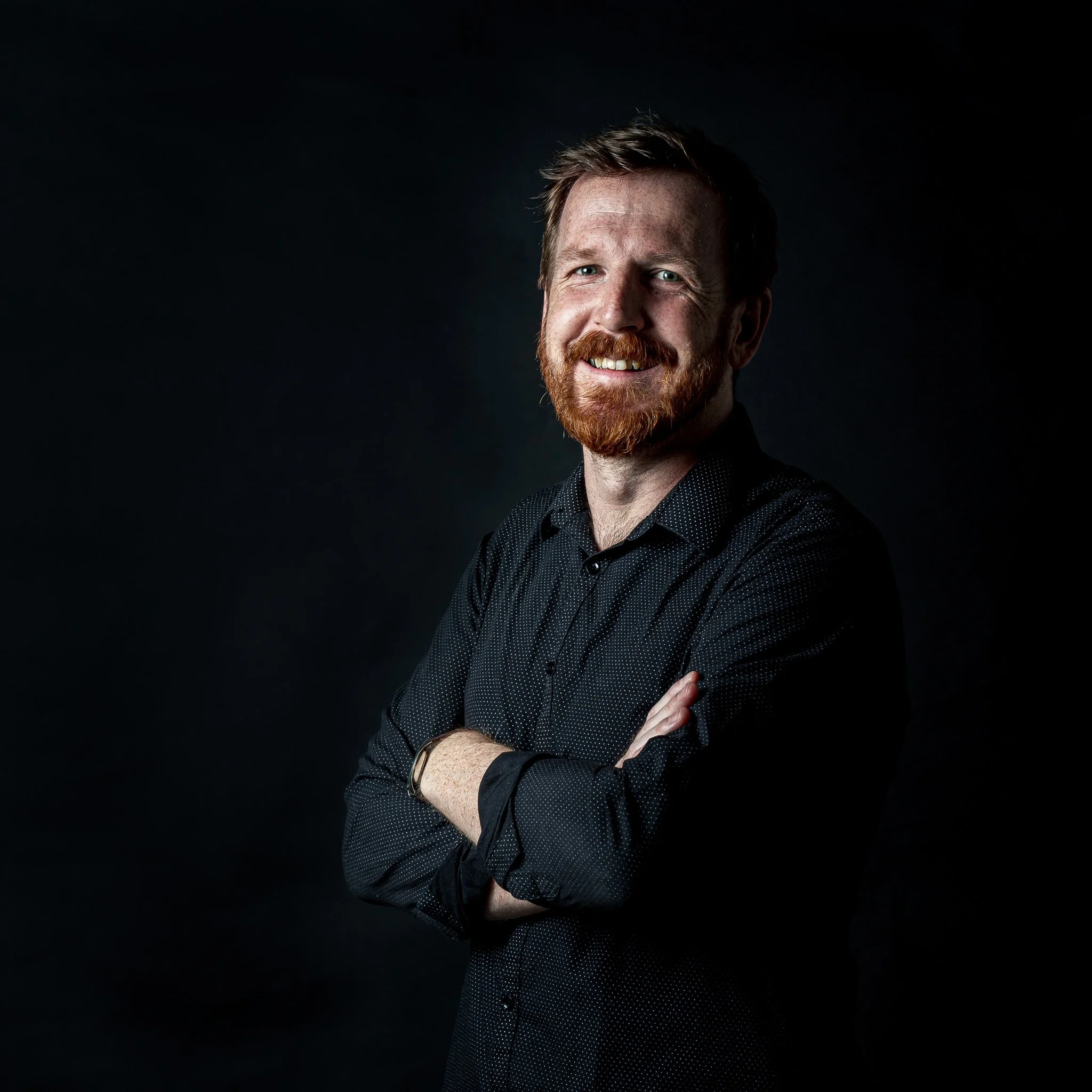 A man with red hair and beard, smiling, standing with arms crossed against a dark background.