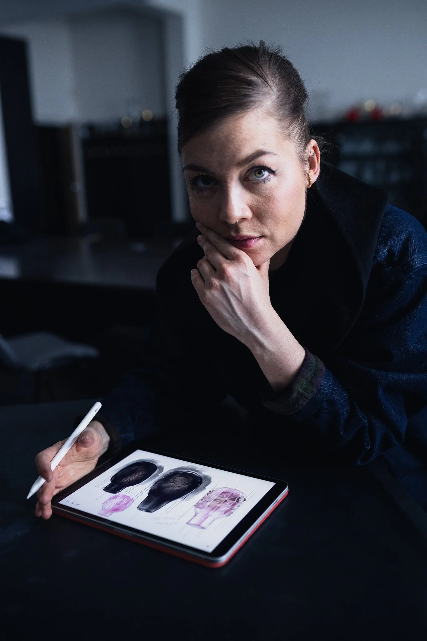 A woman with brown hair and light skin resting her chin on her hand, sitting at a table with a digital drawing tablet displaying sketches of black objects and pink designs, holding a stylus in her right hand.