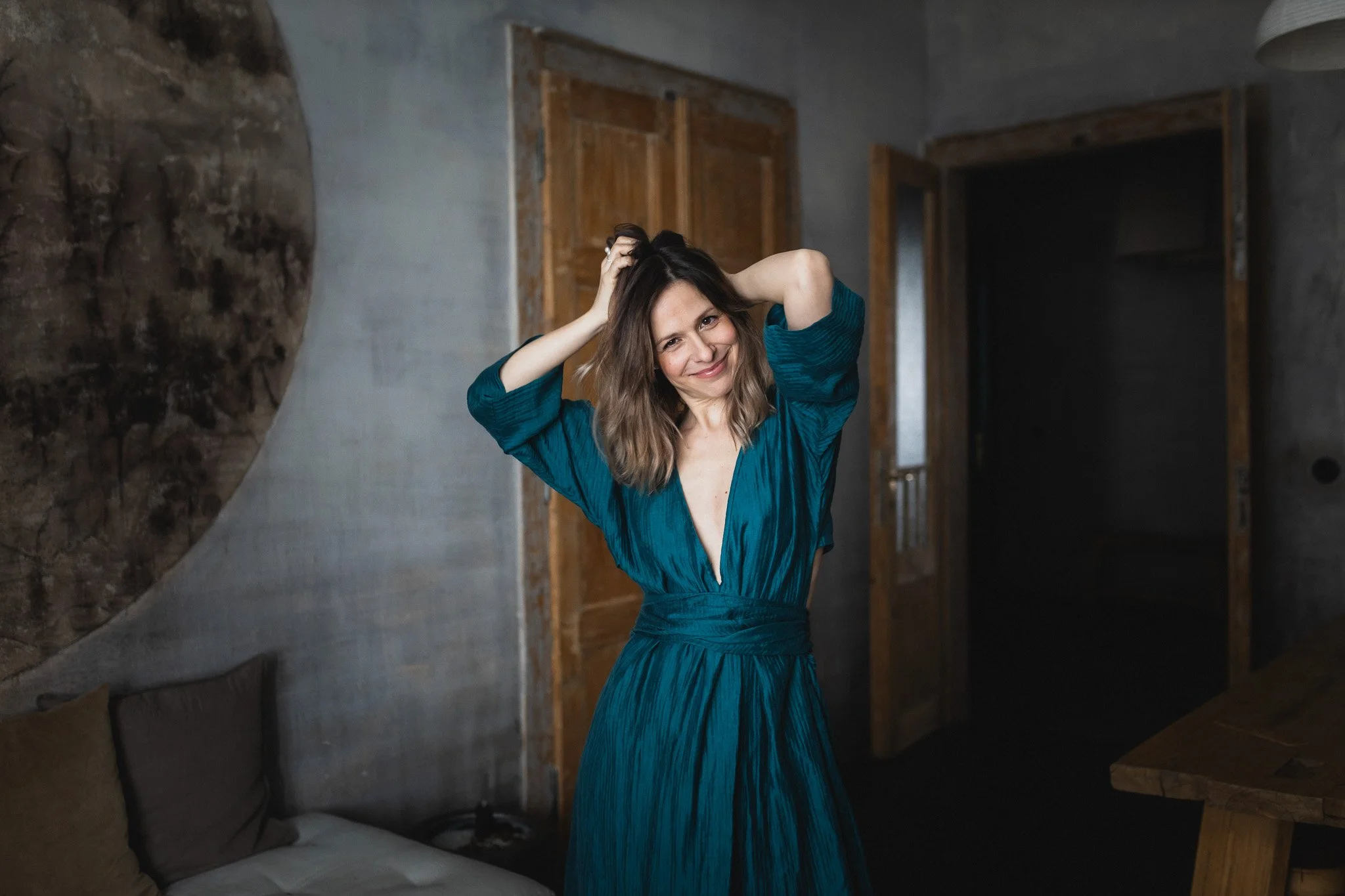 A woman standing in a dimly lit room, smiling, with her hands in her hair, wearing a teal dress with a deep V neckline.