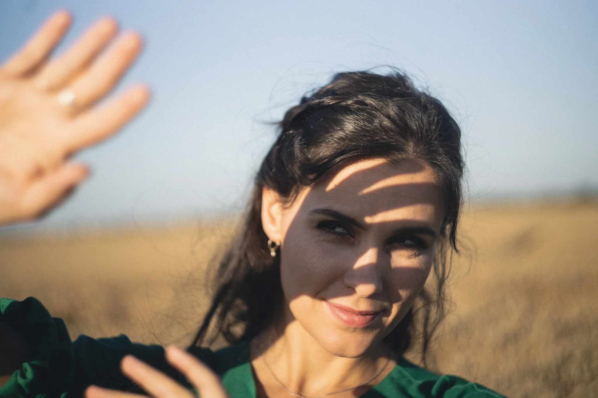 A woman smiling and taking a selfie outdoors, with a shadow in the shape of a hand on her face, in a field under a clear sky.