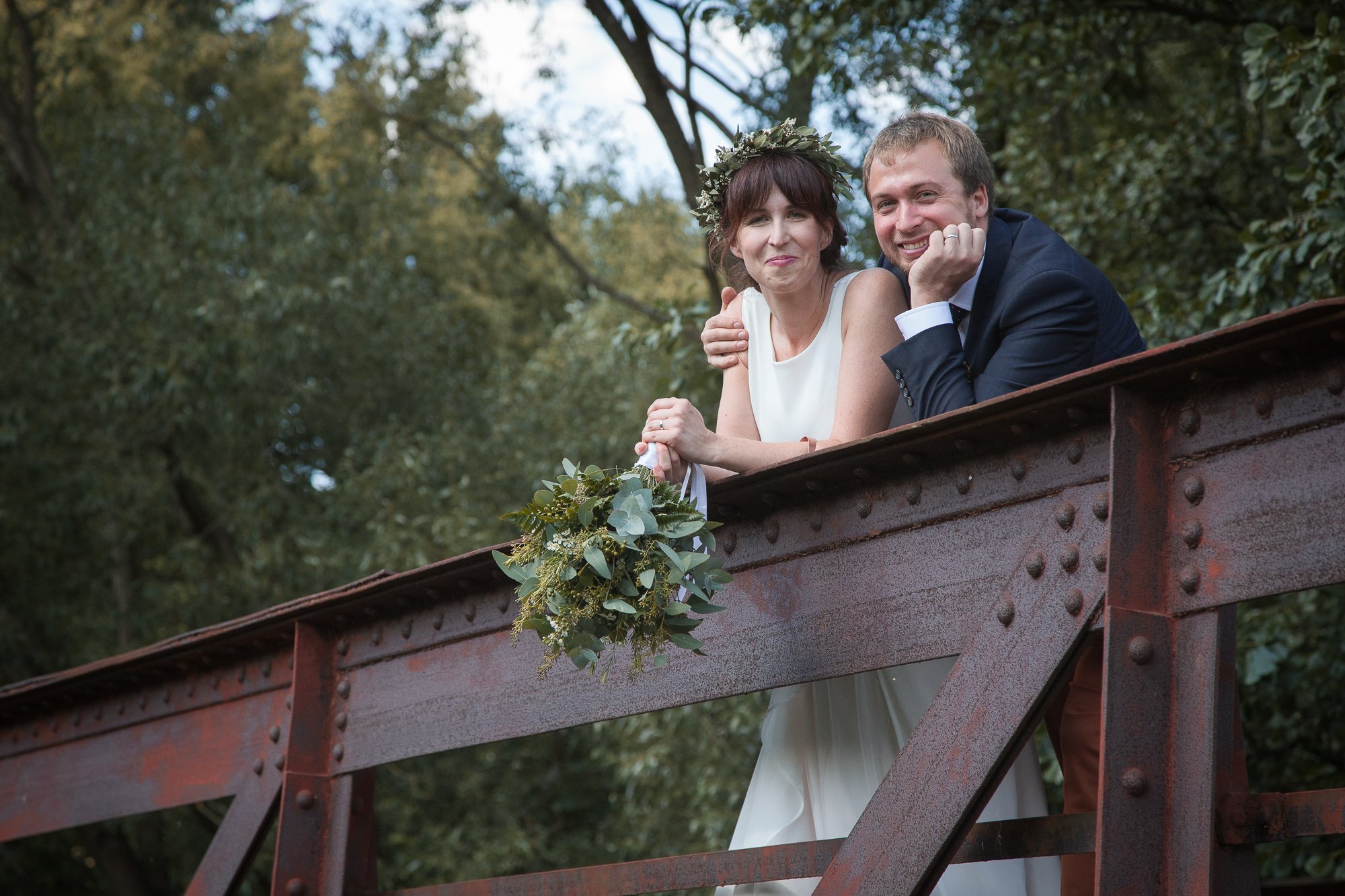 A smiling bride and groom leaning on a rustic bridge railing outdoors, with the bride holding a bouquet of greenery, surrounded by trees.