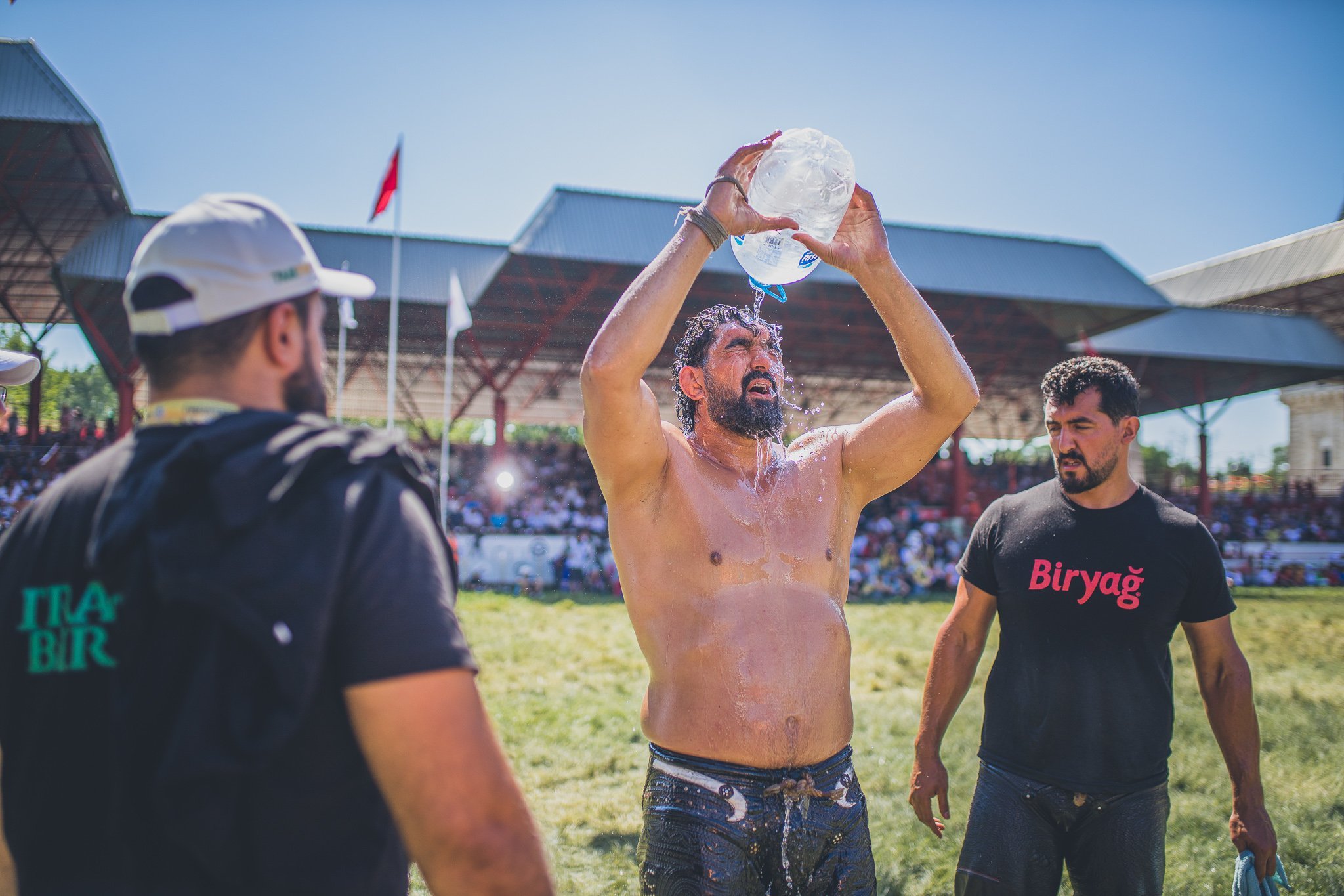 A shirtless man with a beard pouring water over his head during an outdoor event, with two men nearby and a crowd watching in the background.