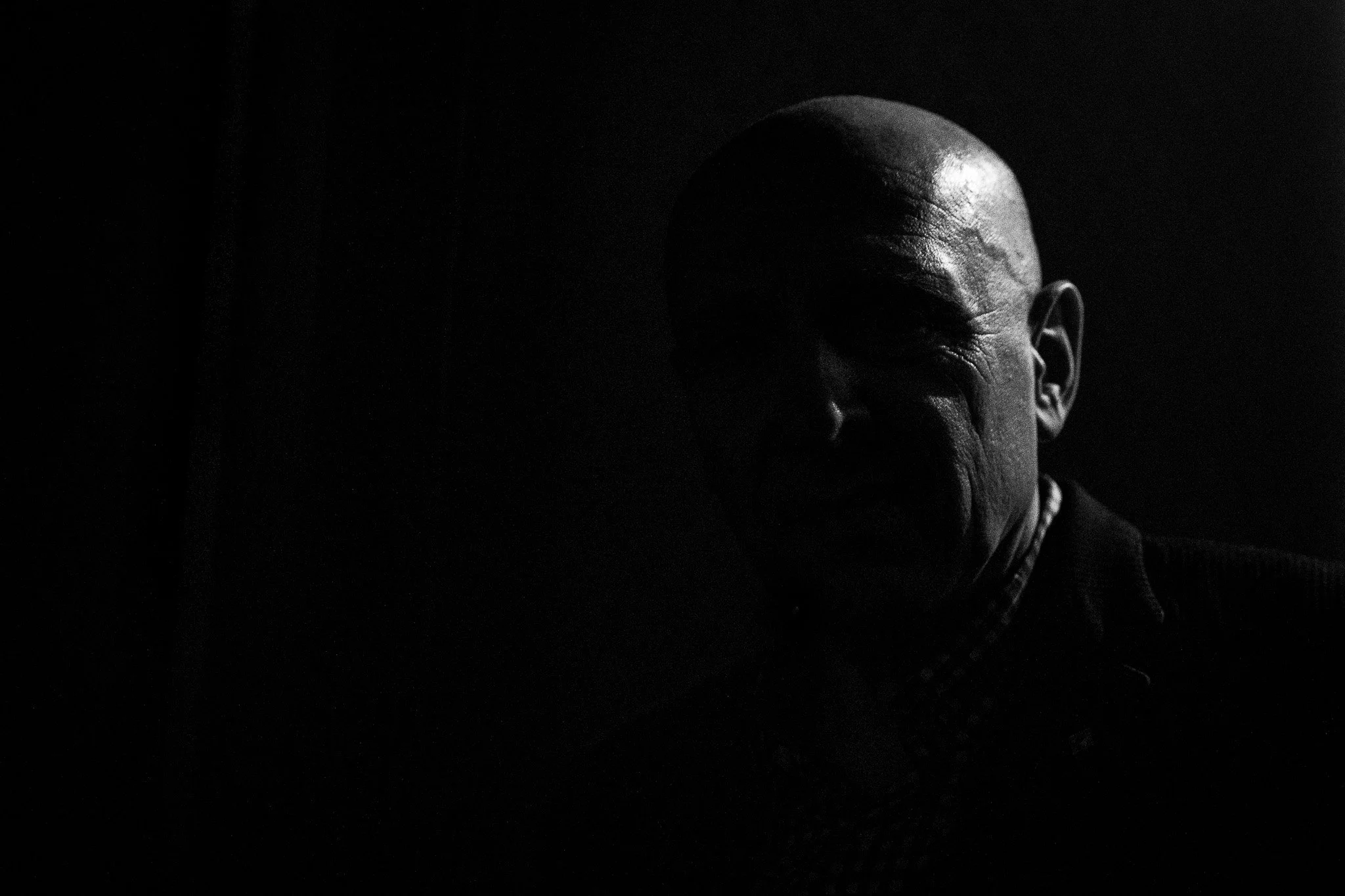 A black-and-white portrait of a bald man in shadow, with a serious expression, looking slightly to the side.