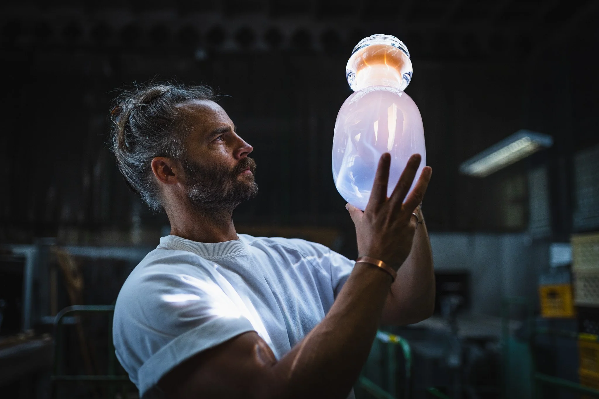 A man with gray hair and a beard, wearing a white shirt, holds a glowing, iridescent glass sculpture indoors, examining it carefully.