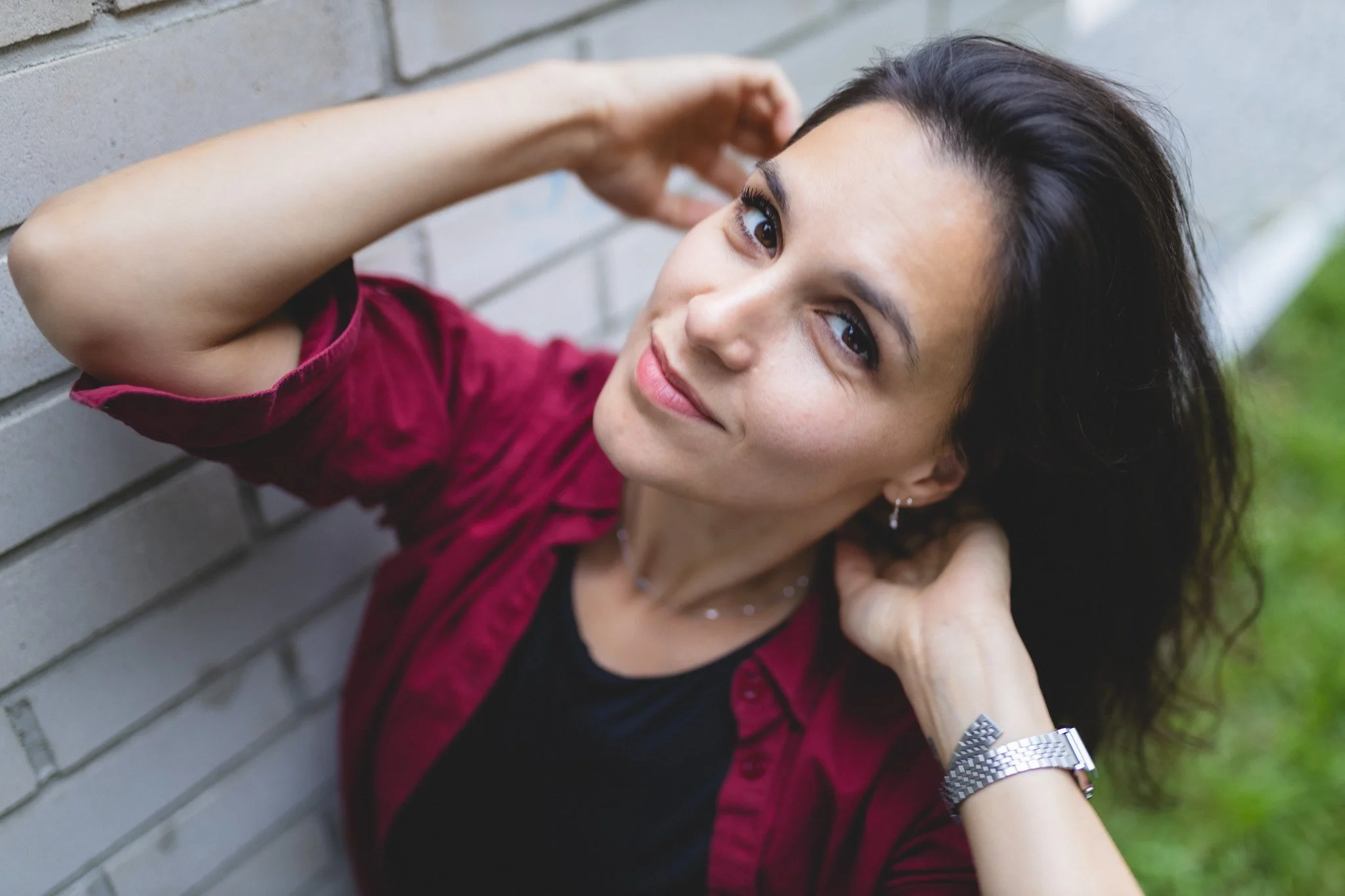 A woman with dark hair and light skin lying against a gray brick wall, looking at the camera with a gentle smile. She is wearing a dark shirt, a red jacket, earrings, and a silver watch.