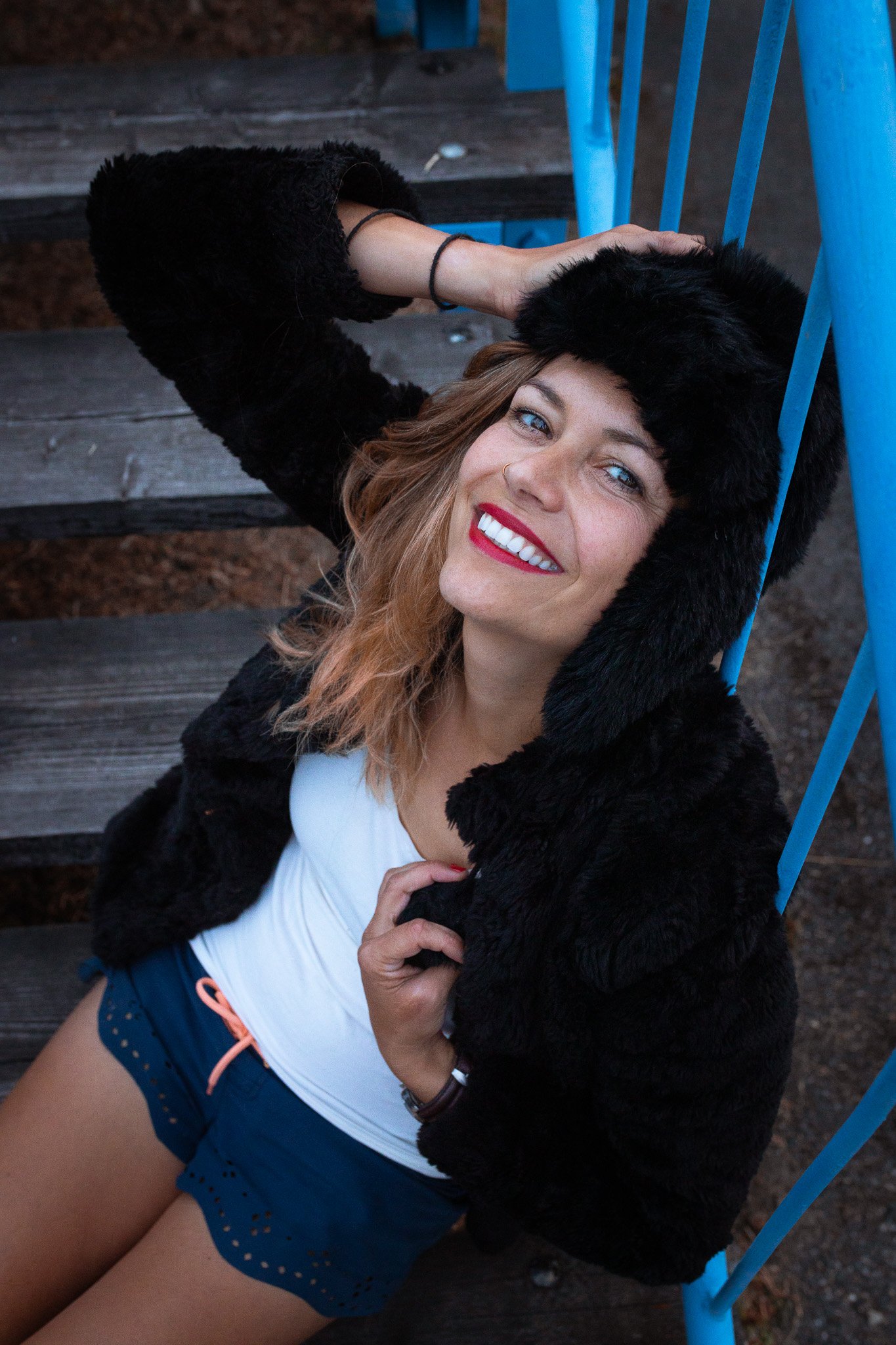 A smiling woman with red lipstick and wavy light brown hair lying on an outdoor wooden staircase, wearing a black faux fur hat and coat, a white t-shirt, and blue shorts, with her hands resting on her coat.