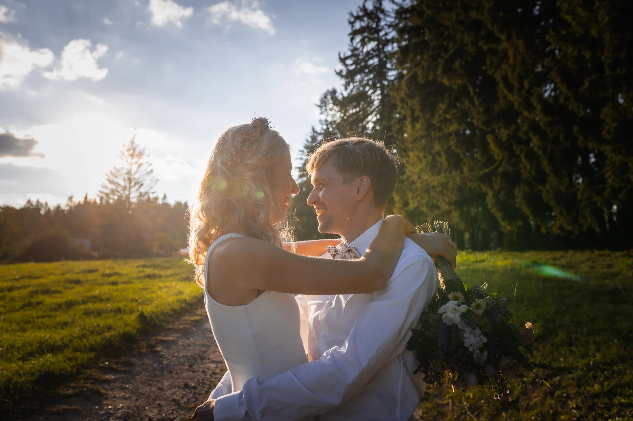 A couple embracing outdoors during sunset, with trees and a grassy field in the background.