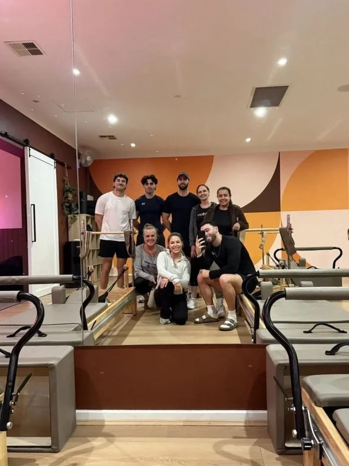 Group of eight people, five women and three men, gathered inside a Pilates studio, posing for a photo in front of Pilates reformer machines.