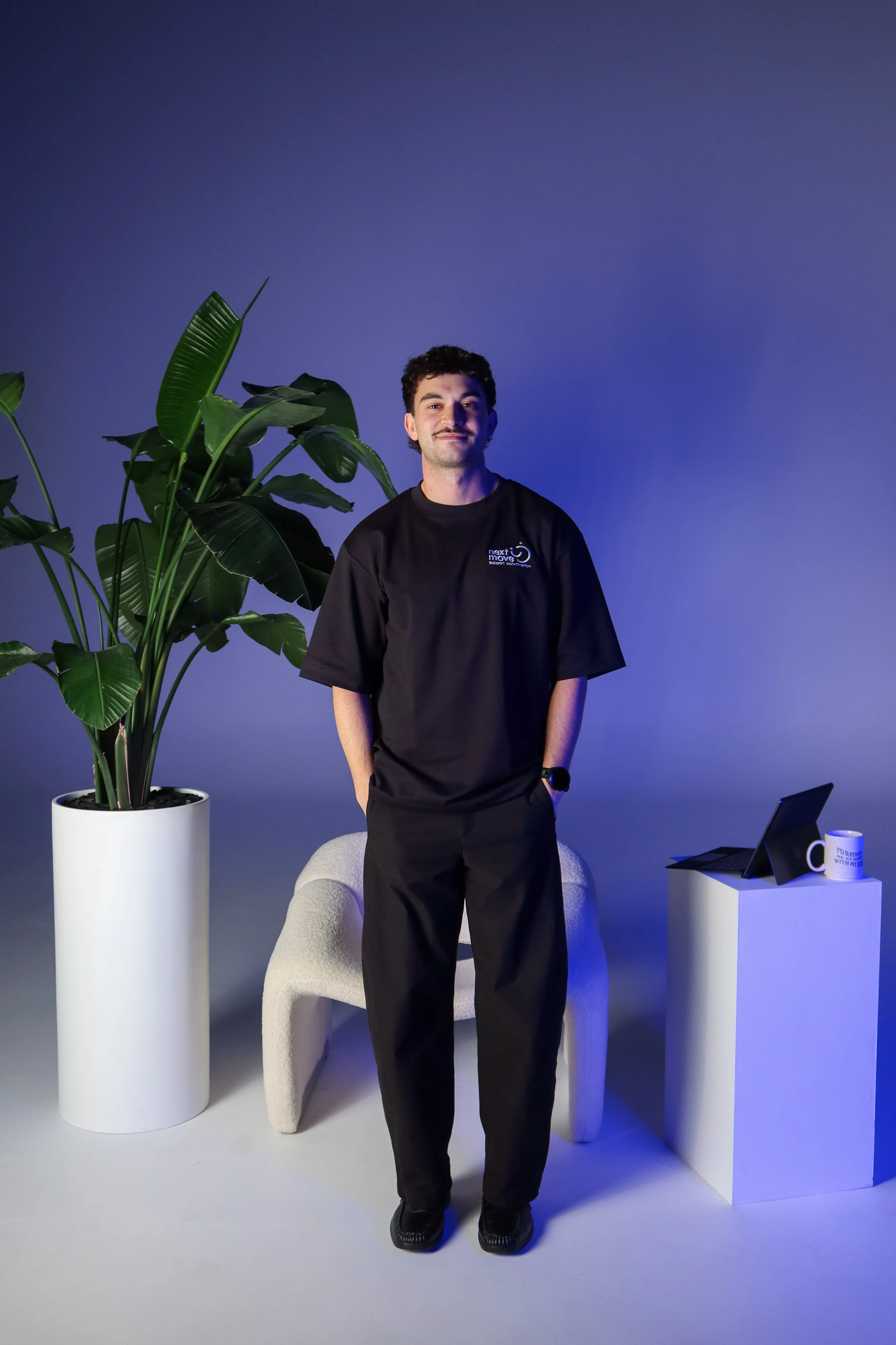 A young man with dark curly hair and a mustache, wearing a black t-shirt with a logo, stands in a modern, minimalistic room with blue ambient lighting. He is in front of a large potted plant, with a white chair behind him, and a white side table holding a laptop and a coffee mug.