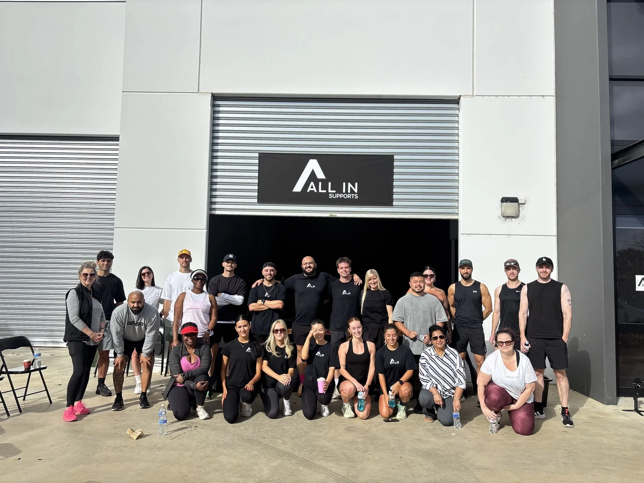 A large group of men and women posing for a photo outside a building with a sign that reads 'ALL IN SUPPORTS'. They are dressed in workout attire, some have water bottles, and a few are smiling or looking at the camera.