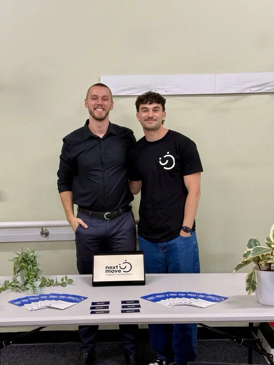 Two men standing behind a table with brochures and a tablet showing the 'next move' logo. The man on the left is in a black button-up shirt, and the man on the right is in a black t-shirt with a smiling face logo. They are smiling and posing for the photo.