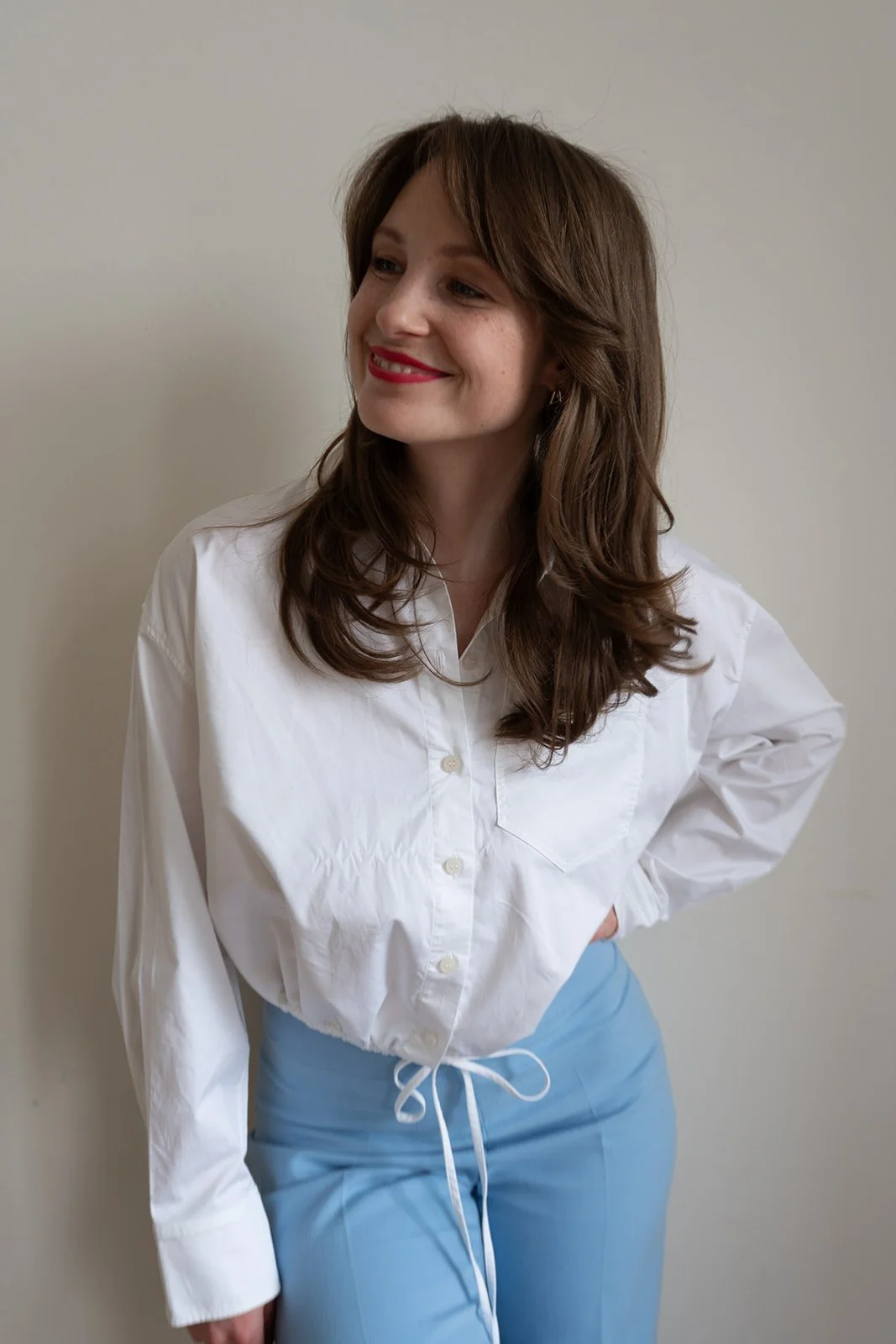 Junge Frau mit braunen, gewellten Haaren, rotem Lippenstift, weißem Hemd und blauer Hose vor einer weißen Wand.