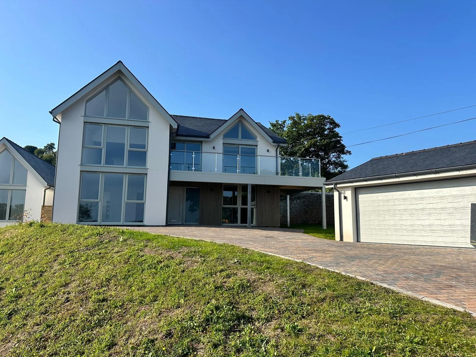 NJPlanning Ltd — A modern, white, multi-story house with large glass windows and a glass balcony, situated on a sloped lawn with a brick driveway, under a clear blue s