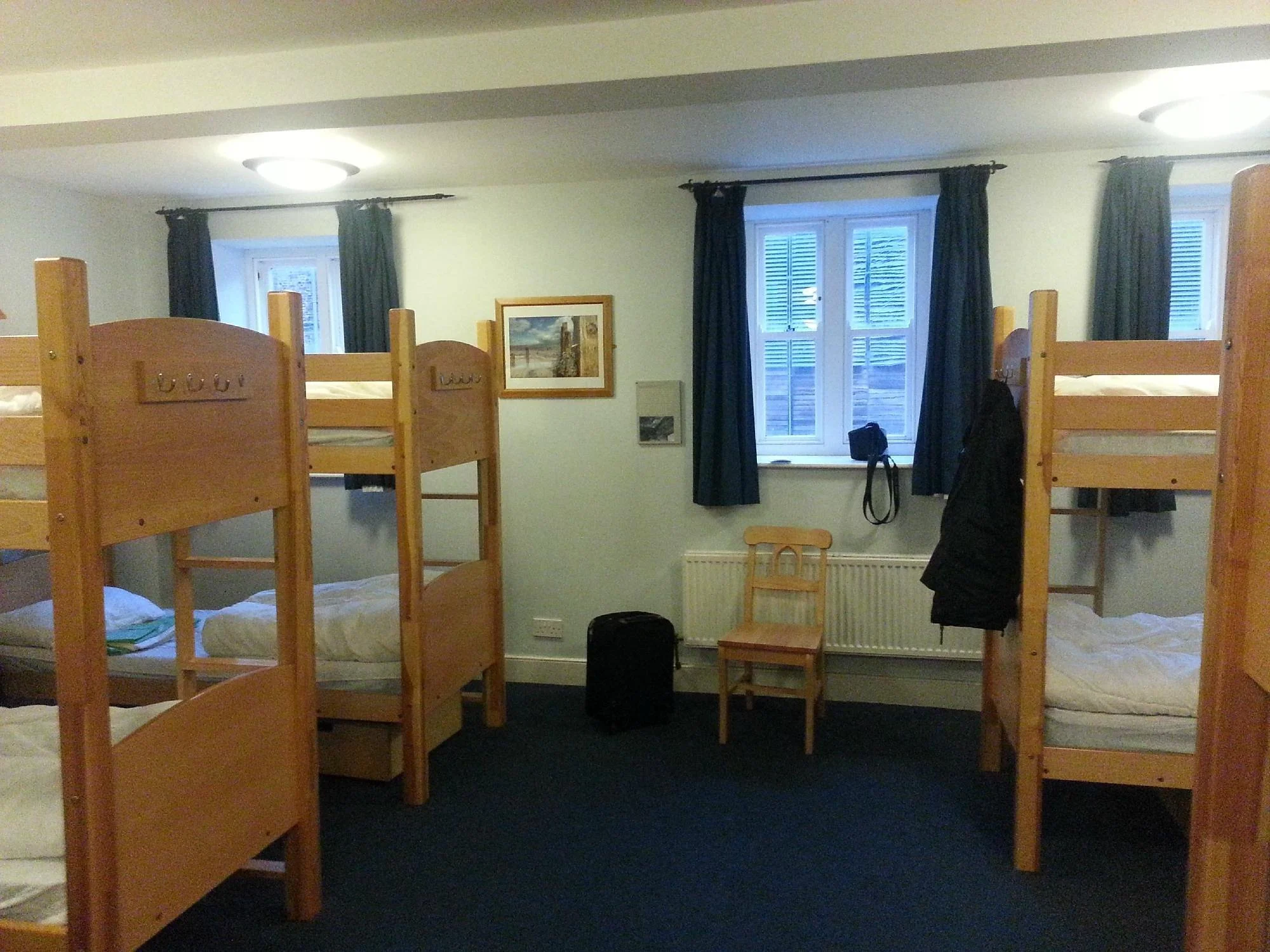 A bedroom with four wooden bunk beds, a window with blue curtains, a painting on the wall, and a chair. There are personal belongings on the beds and at the foot of the beds. The room has two ceiling lights and a dark-colored carpet.