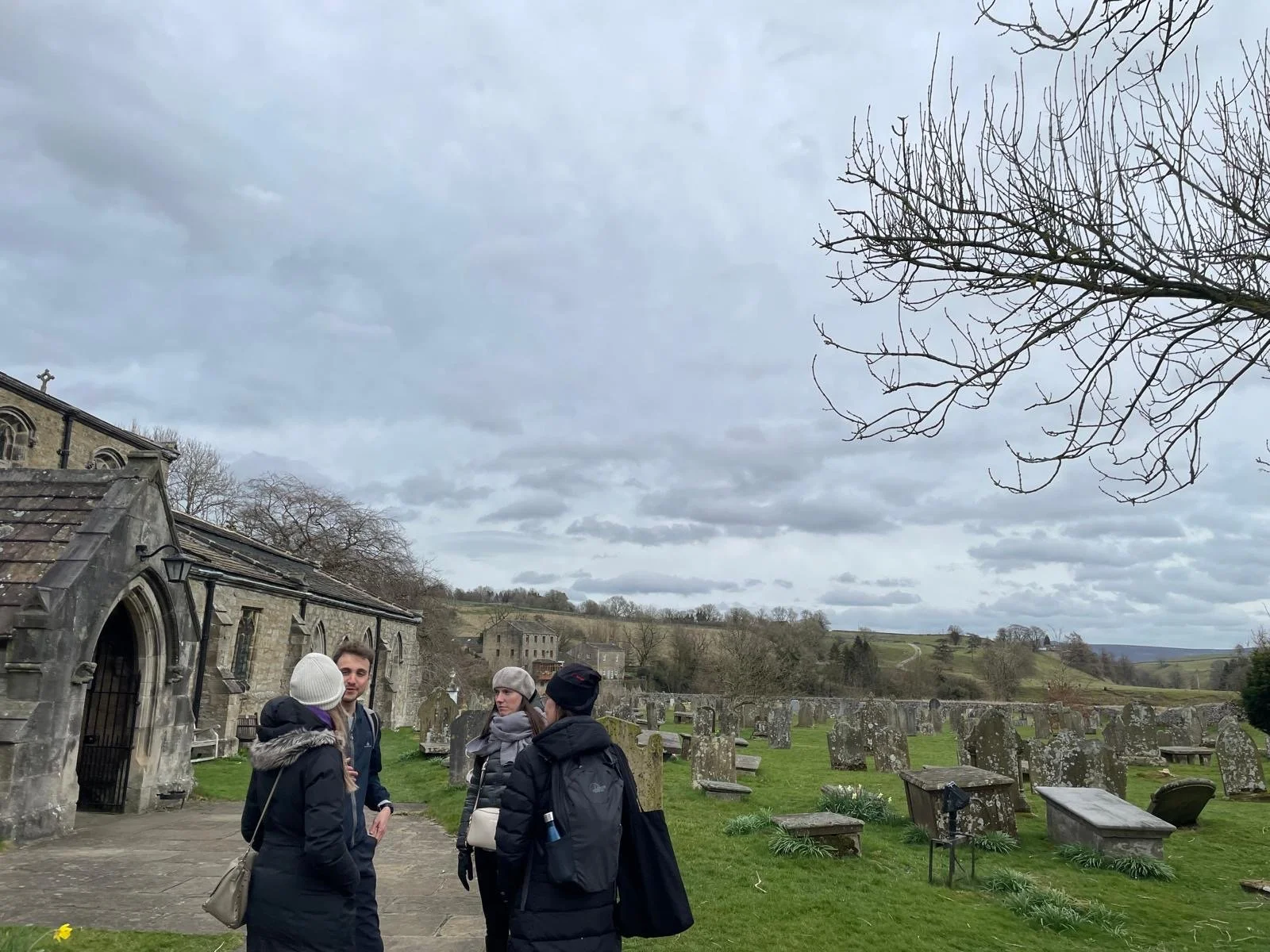 Four people standing near a church and graveyard outdoors on a cloudy day, with rolling hills in the background.