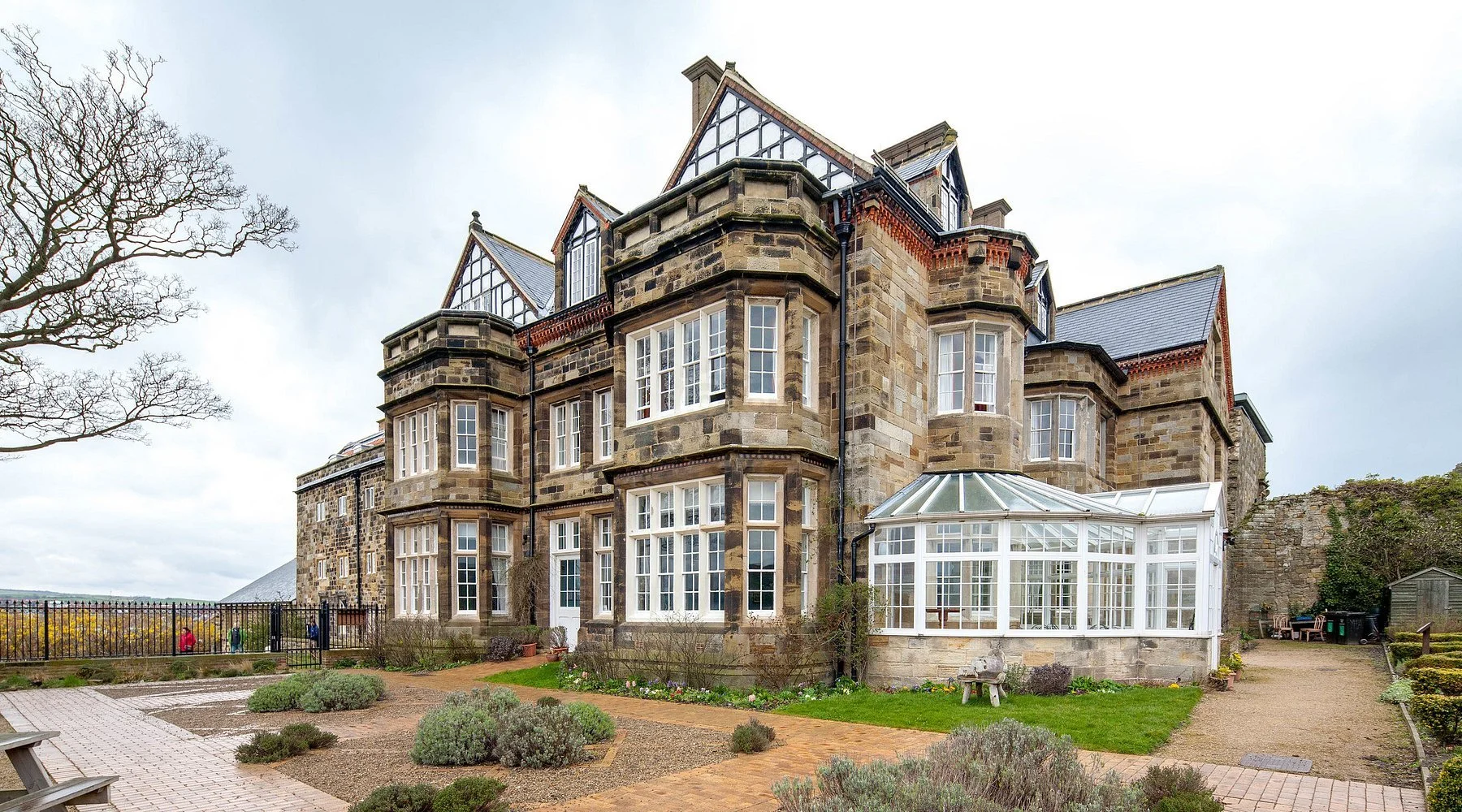 A historic stone mansion with multiple gabled roofs and a glass conservatory attached to the side, set amid landscaped gardens with pathways and shrubs.