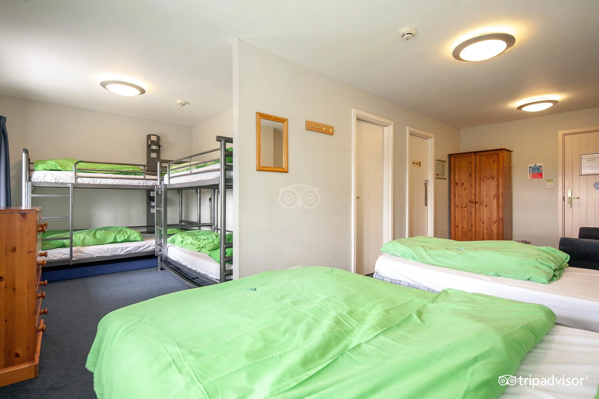 Dormitory room with single and bunk beds, some with green bedding, wooden wardrobe, and white walls with doors and a mirror