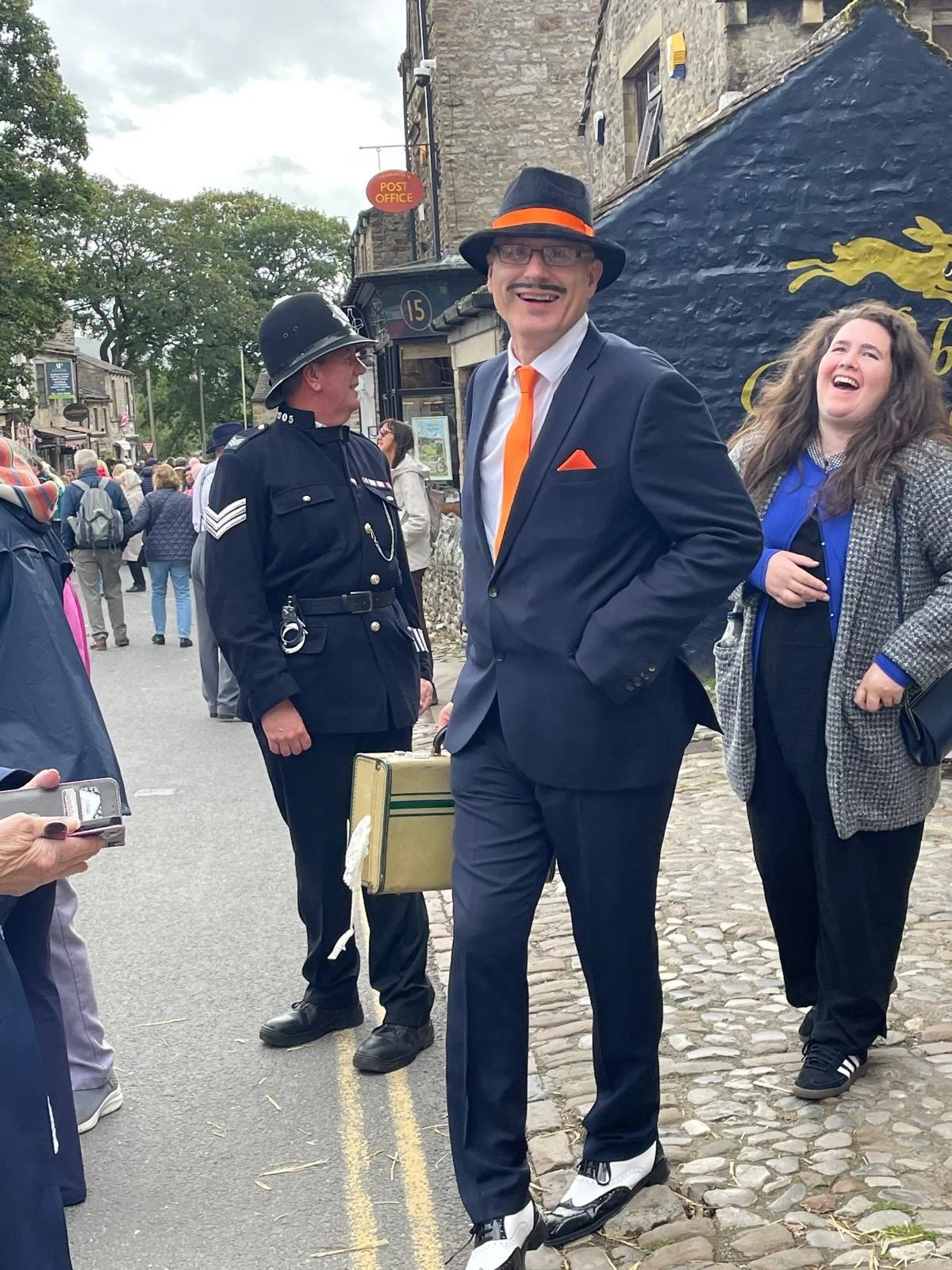 A man in a suit and hat smiling and talking on a busy street with people, including a police officer, around him. The street has cobblestones and historic buildings, with a painted mural in the background.