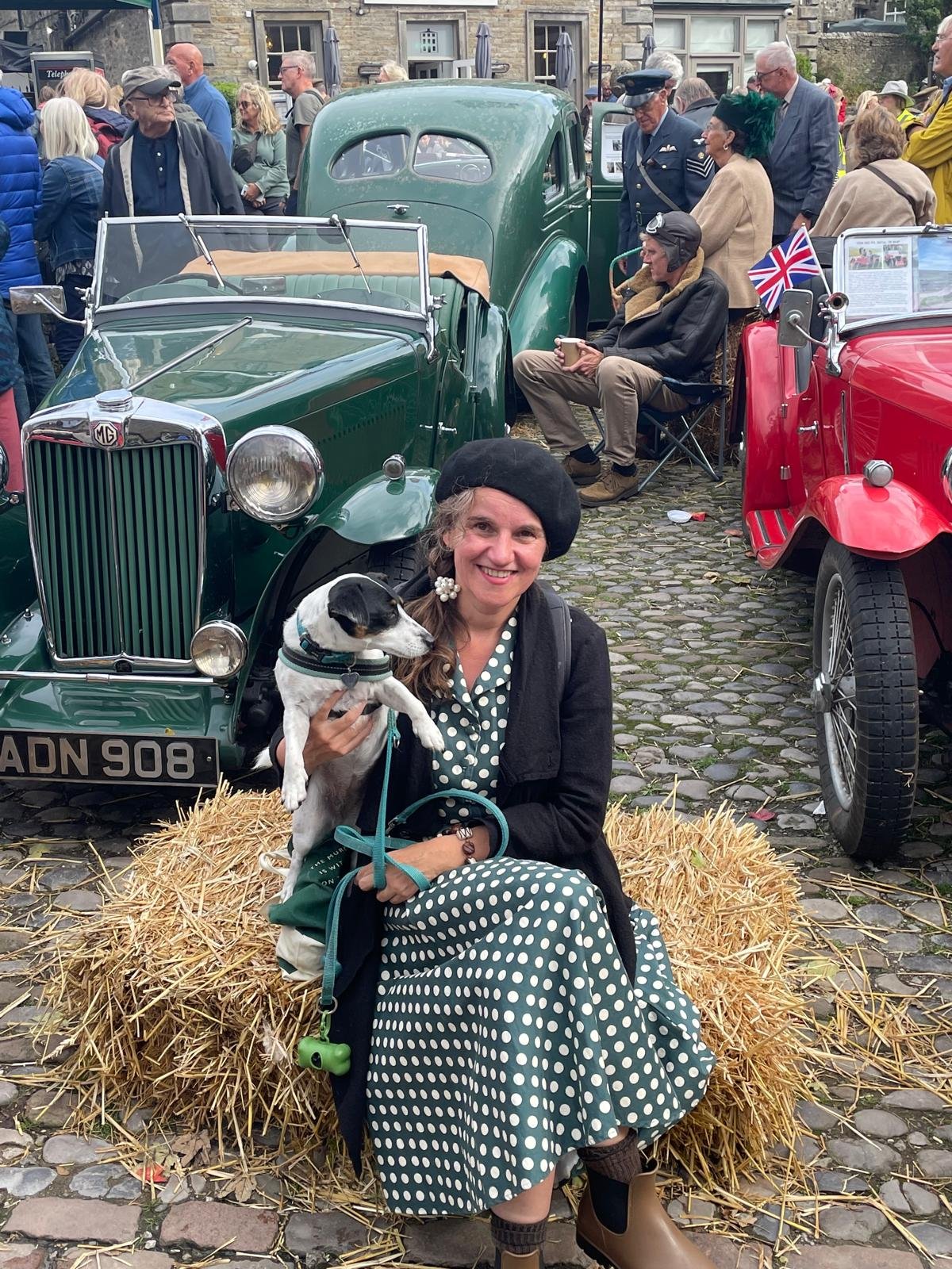 A woman in a polka dot dress and black jacket sitting on a hay bale, holding a small black and white dog, smiling at the camera at a vintage car show. Multiple classic cars and people are seen in the background.