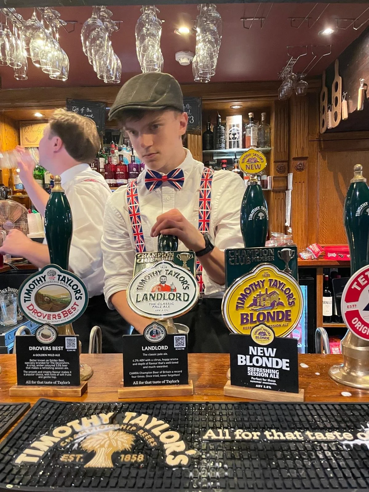 A bartender at a pub wearing a white shirt with a Union Jack bow tie and suspenders, standing behind a bar with various tap handles for Timothy Taylor's beers. The bar has a wooden back with shelves of liquor bottles and hanging glassware. A customer is visible on the left side of the image, and there are promotional signs for different Timothy Taylor's beers on the bar.