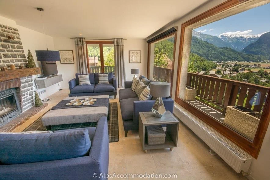 commercial Living room with large window view of mountains and valley, blue and gray sofas, fireplace, small side tables with lamps, and a balcony with outdoor furniture.