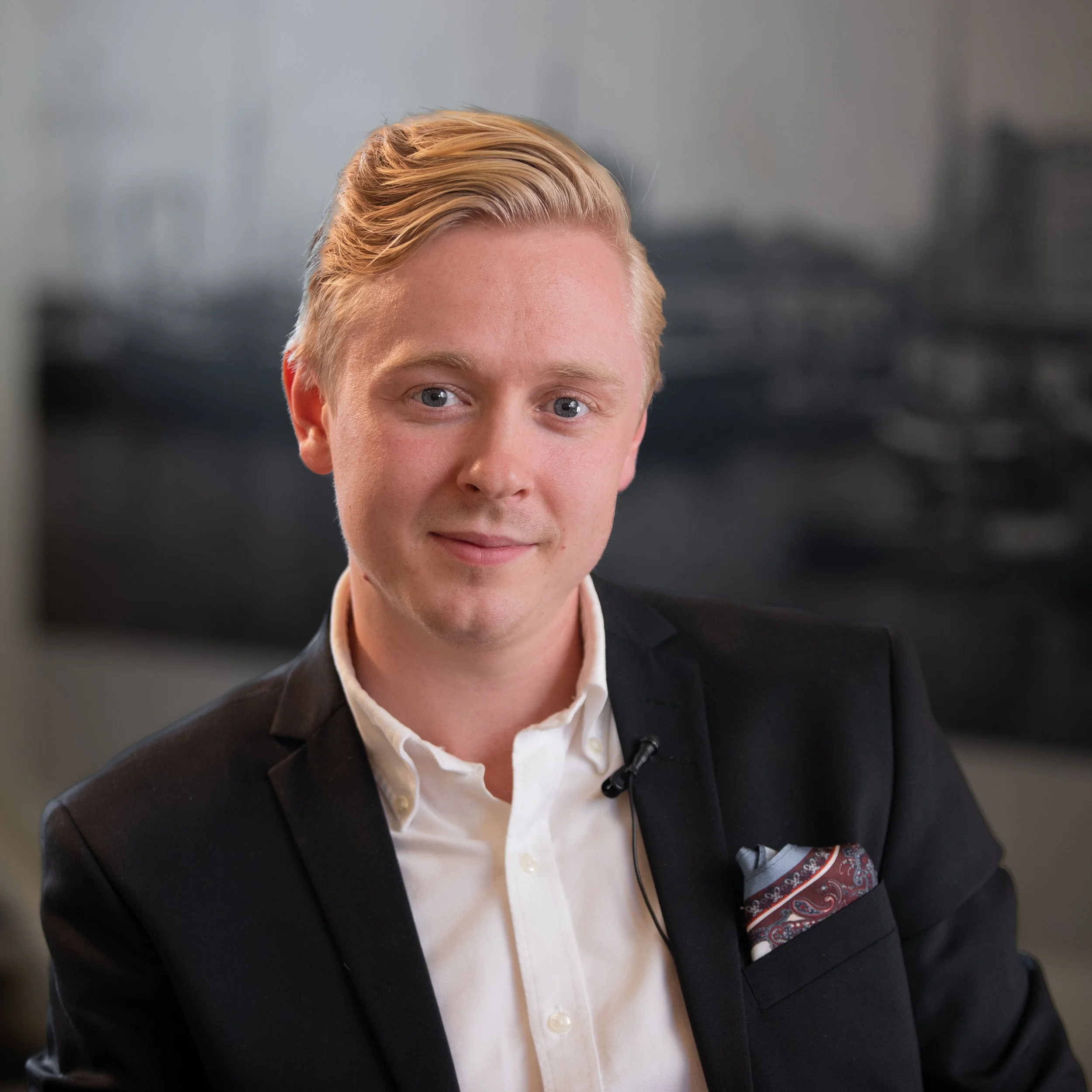 Porträtt av en ung man med blond hår, bär en mörk kostym och vit skjorta, sitter i ett kontorsmiljö.