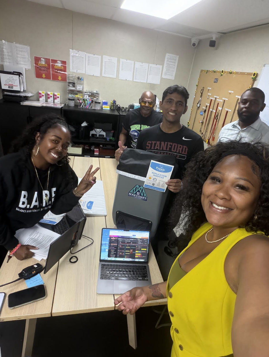 Group of five people in a room with office supplies, smiling at the camera. One woman in yellow taking a selfie, three men standing behind her, and one woman sitting at the table with laptops and documents.