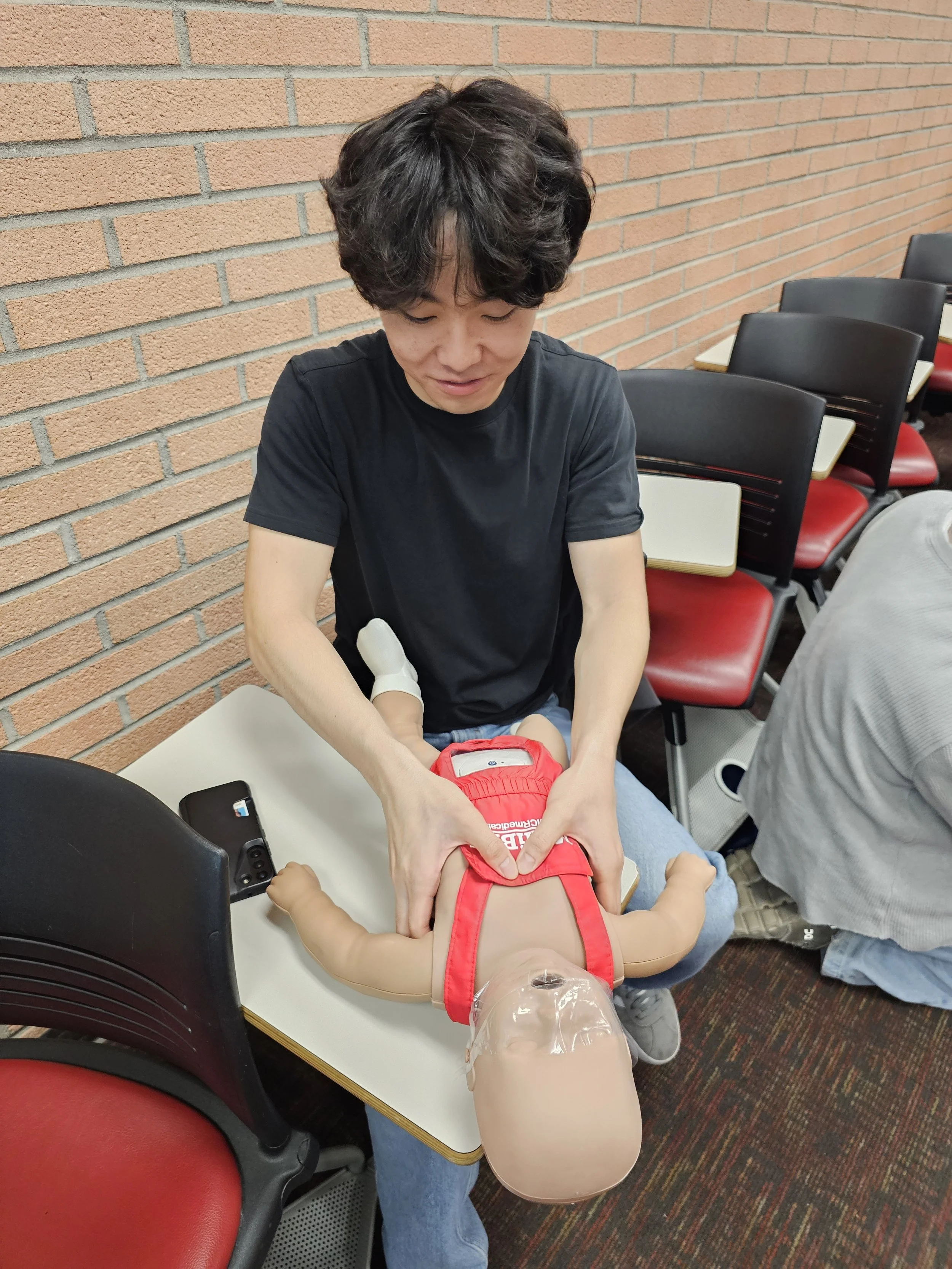 A person with short curly dark hair, wearing a black t-shirt, is administering a vaccination to a small child who is lying on the table, wearing a red harness, inside a room with a brick wall background and chairs.