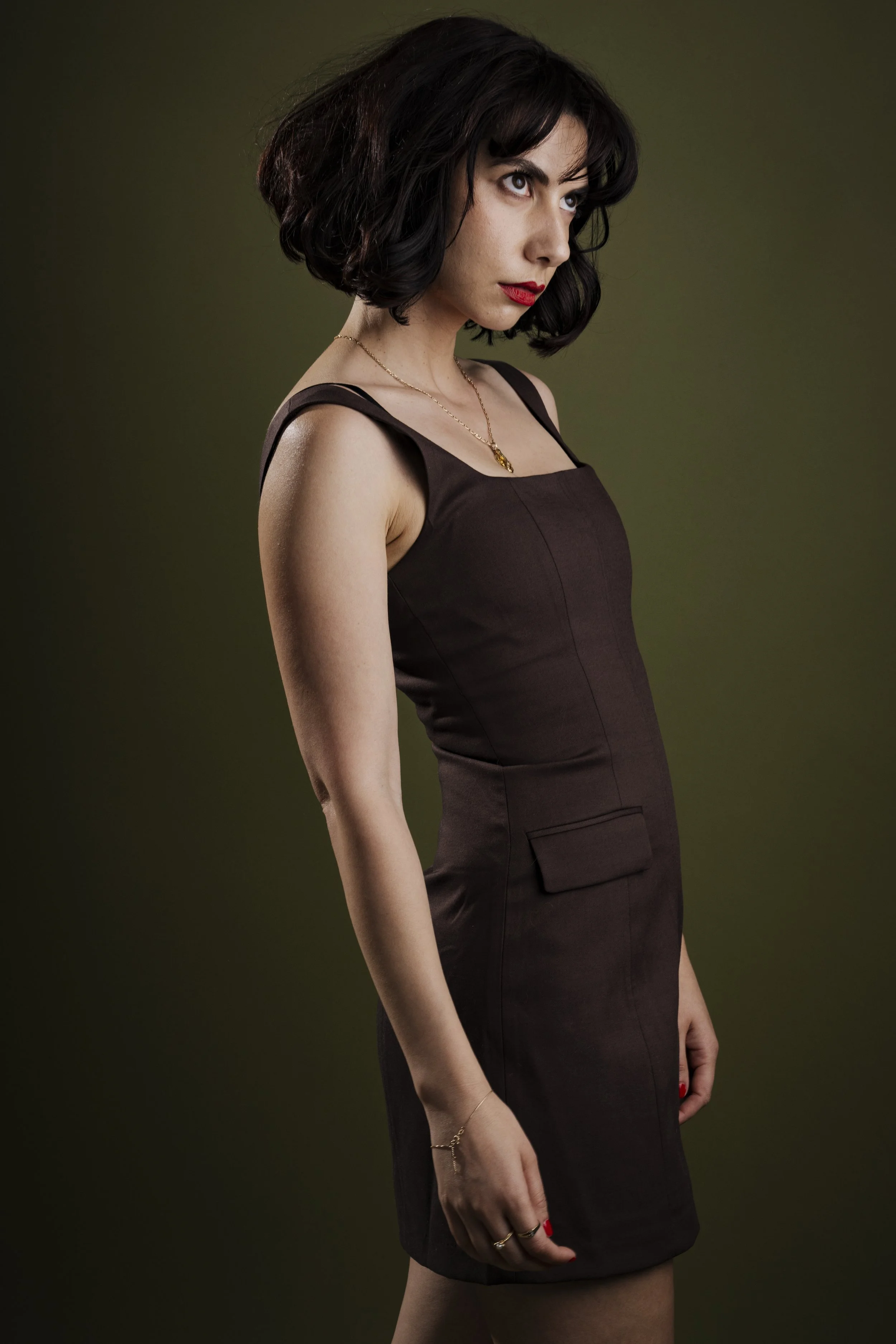 A woman with dark, wavy shoulder-length hair, wearing a dark brown sleeveless dress with pocket details, red lipstick, and minimal jewelry, looking slightly to her right against a dark olive-green background.