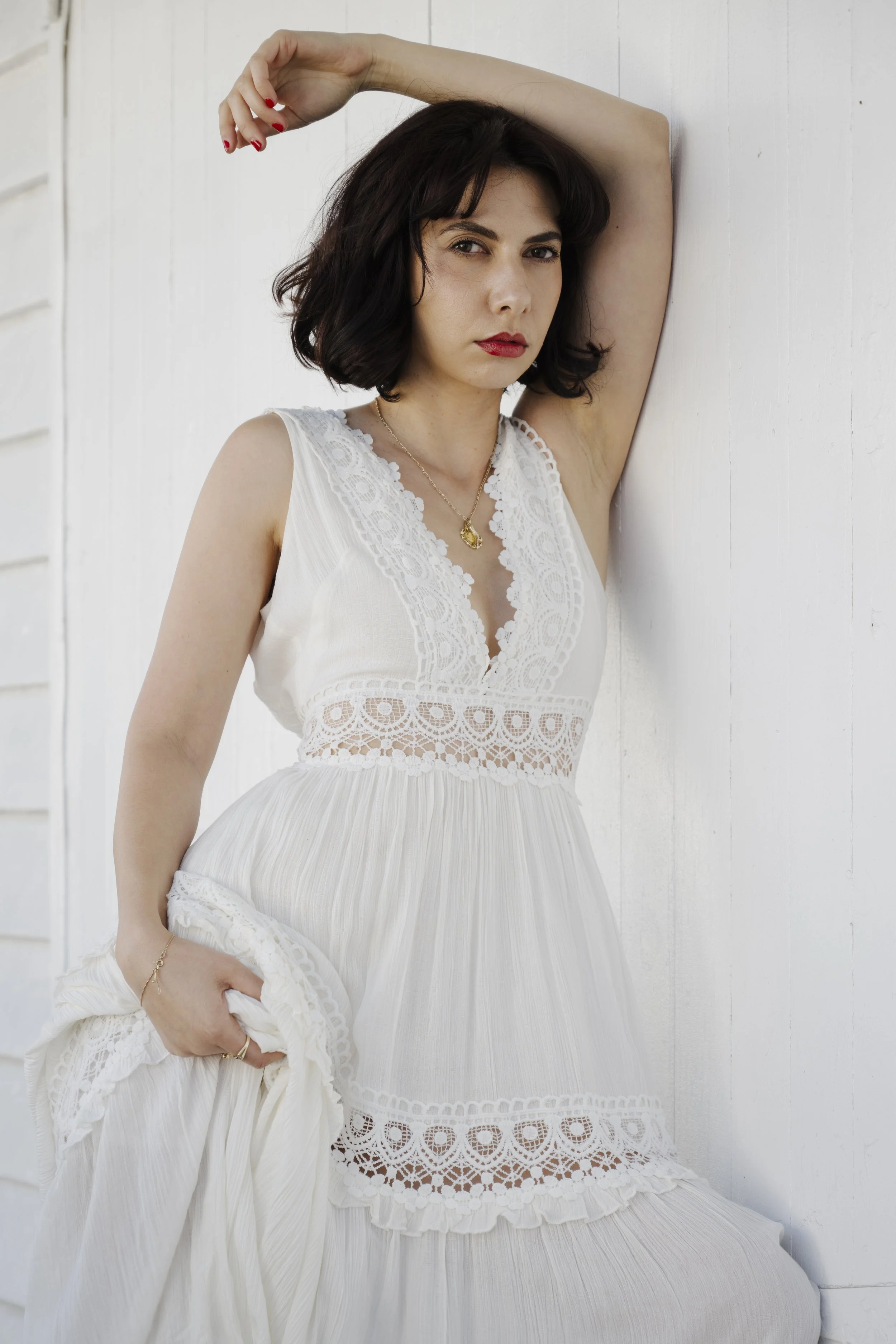A woman in a white dress with lace details leaning against a white wall, with one arm raised and resting on her head, looking directly at the camera.