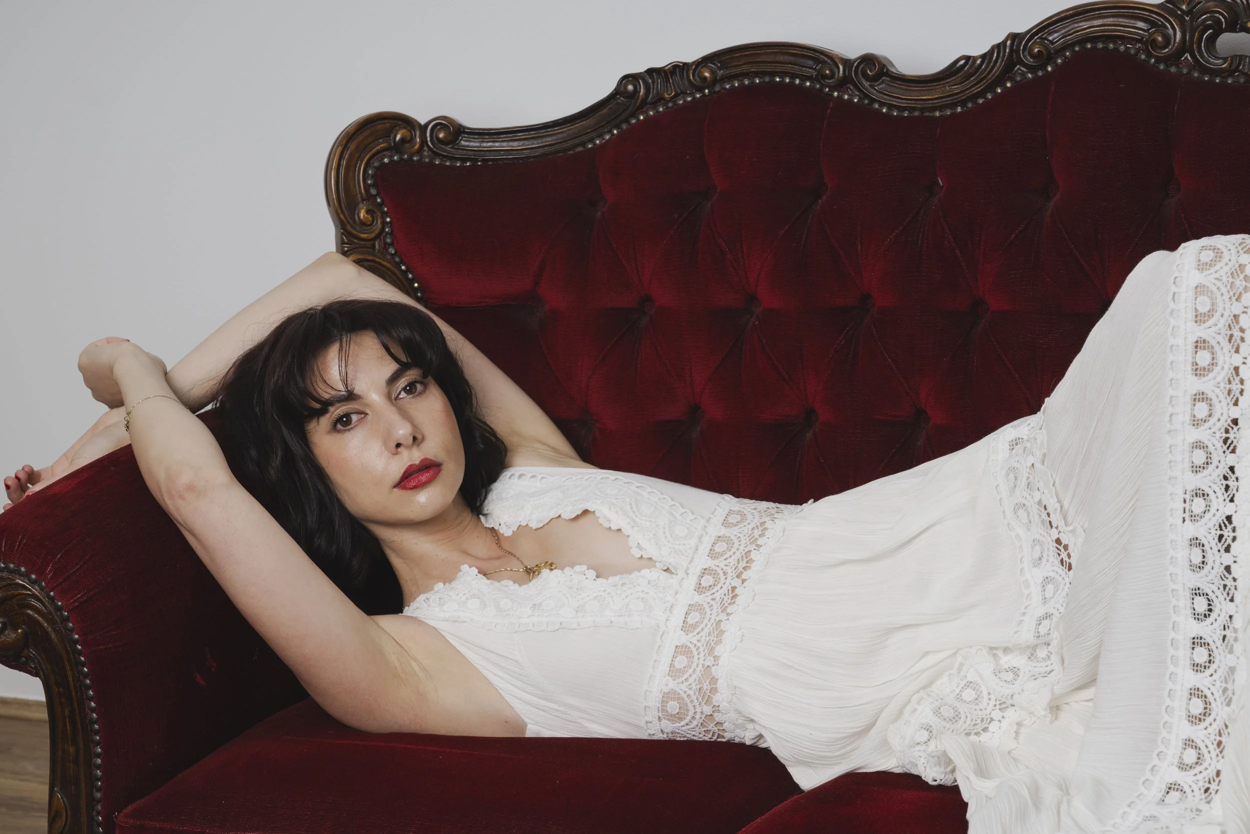 Woman with black hair and red lipstick lying on a vintage red velvet sofa in a white dress.